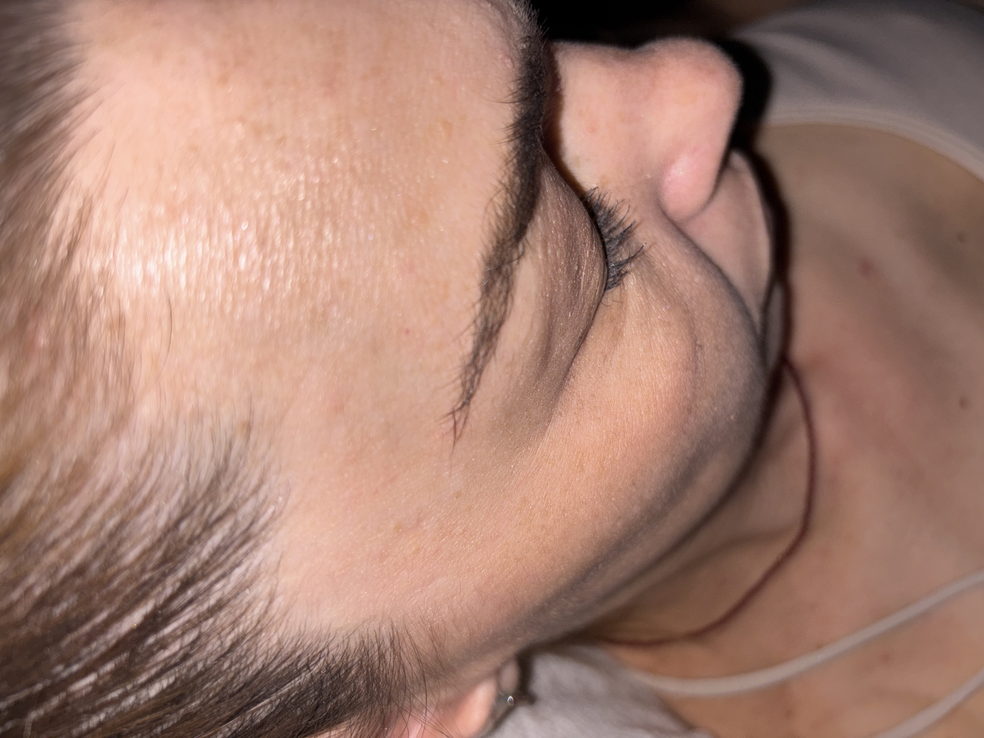 This is a close-up, high-angle shot of a person's face and upper neck, angled downwards. The subject's hair is pulled back, revealing their forehead, eyebrow, eyelashes, eye, and part of their nose and cheek. The skin appears to have some freckles and fine lines. A faint, thin, dark red necklace is visible around the neck. The lighting is dim, with some highlights on the skin. There are no discernible environmental elements that would indicate a specific location within a city.