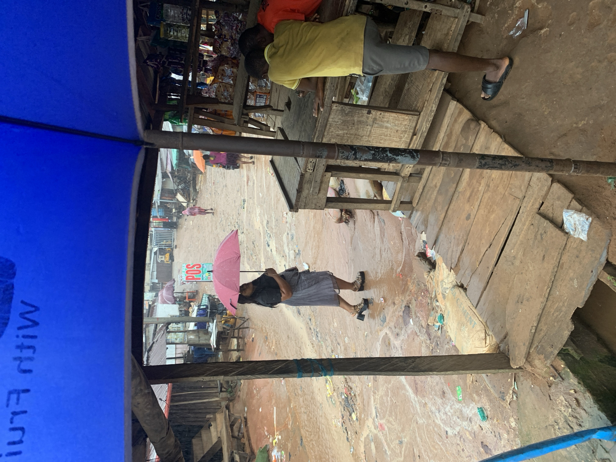 This is a medium-shot, eye-level, outdoor photograph taken on a rainy day in Nkpor, Nigeria. The foreground is dominated by a partially visible blue tarp, likely part of a stall's awning, obscuring the upper portion of the image. A wooden post, weathered and tied with blue string, stands prominently in the left mid-ground.
The environment is a muddy, unpaved street or market area. Puddles of water and debris are visible on the ground. In the mid-ground, a woman walks under a pink umbrella, wearing a black top, a grey skirt, and platform sandals. Behind her, to the left, a person in a pink hooded poncho walks away from the camera. Further back, an orange umbrella is visible. A stall structure with a "POS" sign is present.
In the right mid-ground, two young men are seen near a wooden stall. One, wearing a yellow t-shirt and grey shorts, leans on the stall, and the other, wearing an orange shirt, stands behind him. The stall displays various packaged goods.
The background shows more market structures, some with corrugated metal roofs, and a yellow tricycle, suggesting a bustling urban environment. The overall impression is one of a busy, somewhat dilapidated, open-air market during inclement weather.