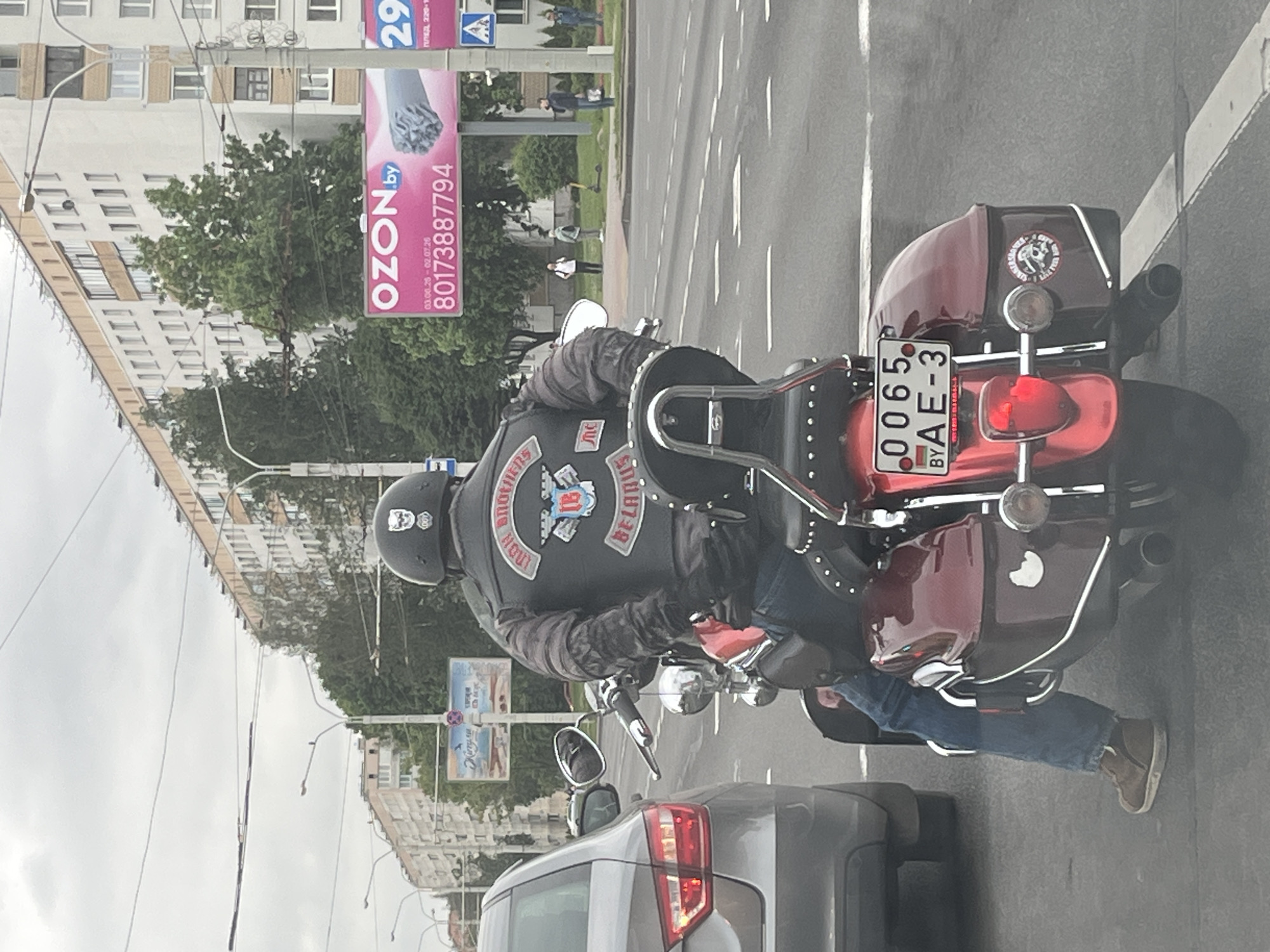 An overhead shot captures a biker on a red motorcycle, likely stopped in traffic or at an intersection, in Minsk, Belarus. The biker wears a black leather jacket adorned with patches, including "BROTHERS" as a top rocker, "BELARUS" as a bottom rocker, and a central emblem featuring a stylized 'B'. A small 'MC' patch is also visible. The biker wears a black helmet with a skull-like motif, blue jeans, and brown boots.
The motorcycle is a cruiser-style bike with chrome accents and saddlebags. Its Belarusian license plate reads "0065 BY AE-3", with "BELARUS" and "Minsk" printed below the numbers. A sticker depicting a skull and text, possibly "UNITED WE STAND," is visible on the rear saddlebag.
In the background, tall, light-colored apartment buildings line the street, partially obscured by trees. A large pink advertising banner for "OZON.by" is prominently displayed, showing a phone number "80173887794" and dates "03.06.20 - 02.07.20". Other people can be seen walking on the sidewalk. The sky is overcast, suggesting daytime. Part of a grey car is visible in the immediate foreground.