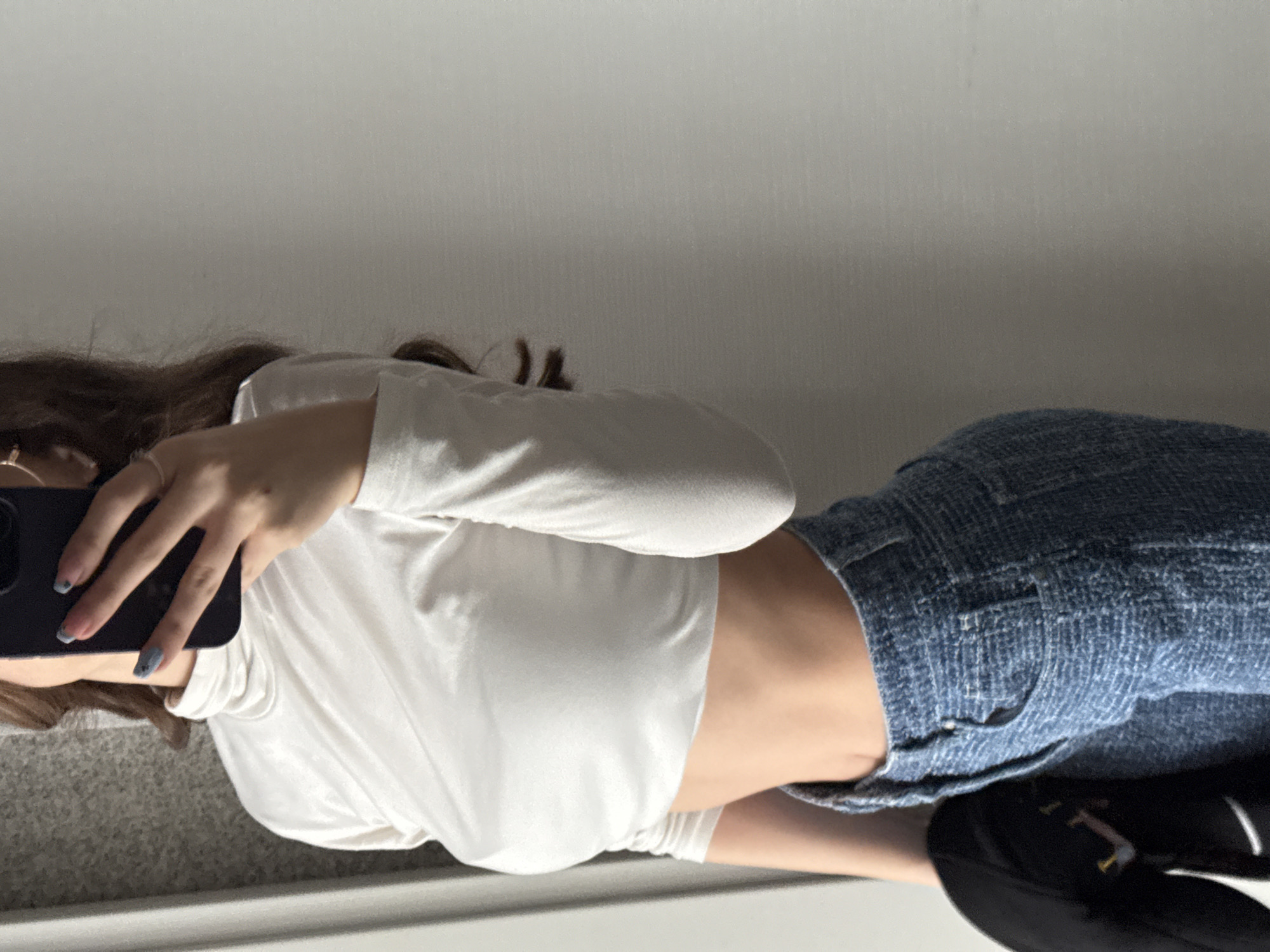 A person is visible from the shoulders to the thighs, wearing a long-sleeved white top and blue denim shorts. The person is holding a smartphone in their left hand, presumably taking a selfie. A black sneaker is visible in the lower right corner. The background is a plain wall. The location is not identifiable as Sofia, Bulgaria from this image.
