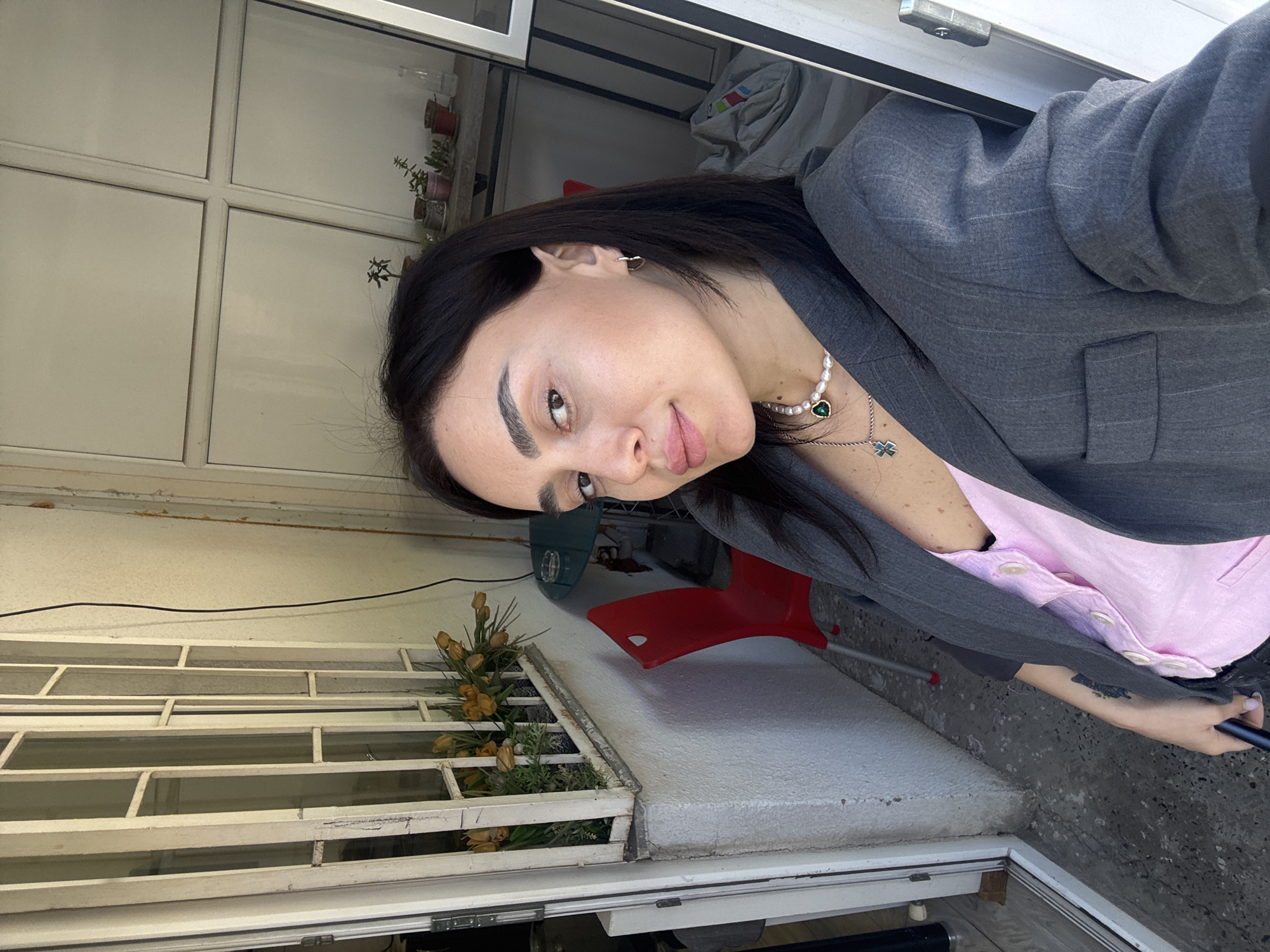 A woman with dark hair and defined eyebrows, wearing a gray blazer over a pink shirt and a pearl necklace with a green pendant, is taking a selfie. She is facing slightly to the right and looking at the camera. The background includes a white-framed window with multiple panes, some plants in a window box, and a red plastic chair. The location appears to be an exterior space, possibly a balcony or patio. Based on the architecture and general aesthetic, the location is likely in the city of Tbilisi, Georgia.