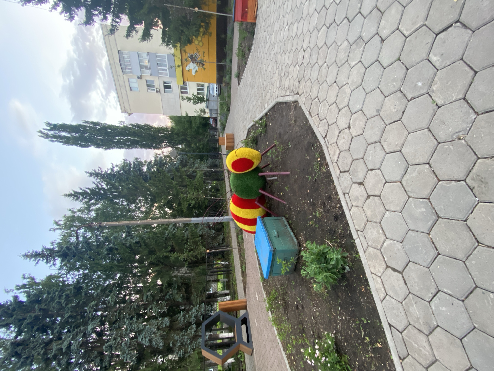 The image captures a whimsical outdoor art installation in the city of Ufa, Russia, featuring a large, colorful insect sculpture with segments of red, yellow, and green, resting in a garden bed. To the left of the insect, a green wooden box with a blue lid is placed, possibly a planter or storage. Adjacent to the garden bed is a hexagonal bench, crafted from wood and metal. In the background, a multi-story residential building stands, adorned with a large mural depicting a bee and a honeycomb pattern. The scene is further enhanced by lush green trees and a paved walkway made of hexagonal tiles that curves through the area. The sky above is partly cloudy, suggesting either dawn or dusk.