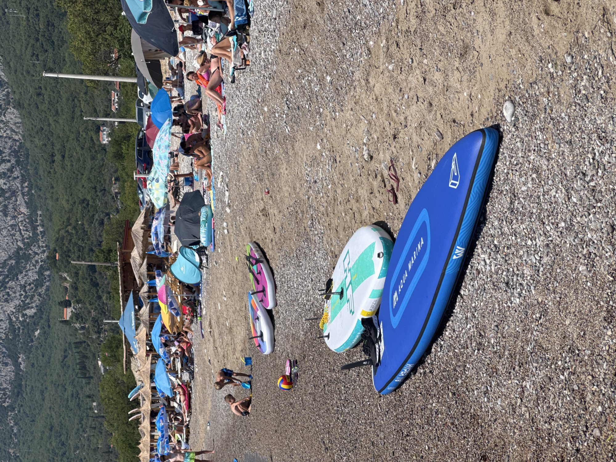 The image depicts a crowded beach in Đurovići, Montenegro, under a clear sky. The beach is covered with pebbles and sand. In the foreground, two inflatable paddleboards are laid out on the sand; one is blue with the brand "Aqua Marina" visible, and the other is white and teal with green accents. Nearby, a smaller white and teal inflatable paddleboard lies on its side, alongside a yellow and red beach ball and a pair of flip-flops.
Further back, several beach umbrellas and straw shelters provide shade for sunbathers. Many people are seen lounging on chairs and towels, some are partially visible, while others are obscured by umbrellas. Children are playing on the beach; two young boys are near the water's edge, one digging in the sand with a shovel. The background shows a dense green hillside with some buildings visible amongst the trees, and a rocky mountain range looms behind it. The sea is not clearly visible in this particular shot.