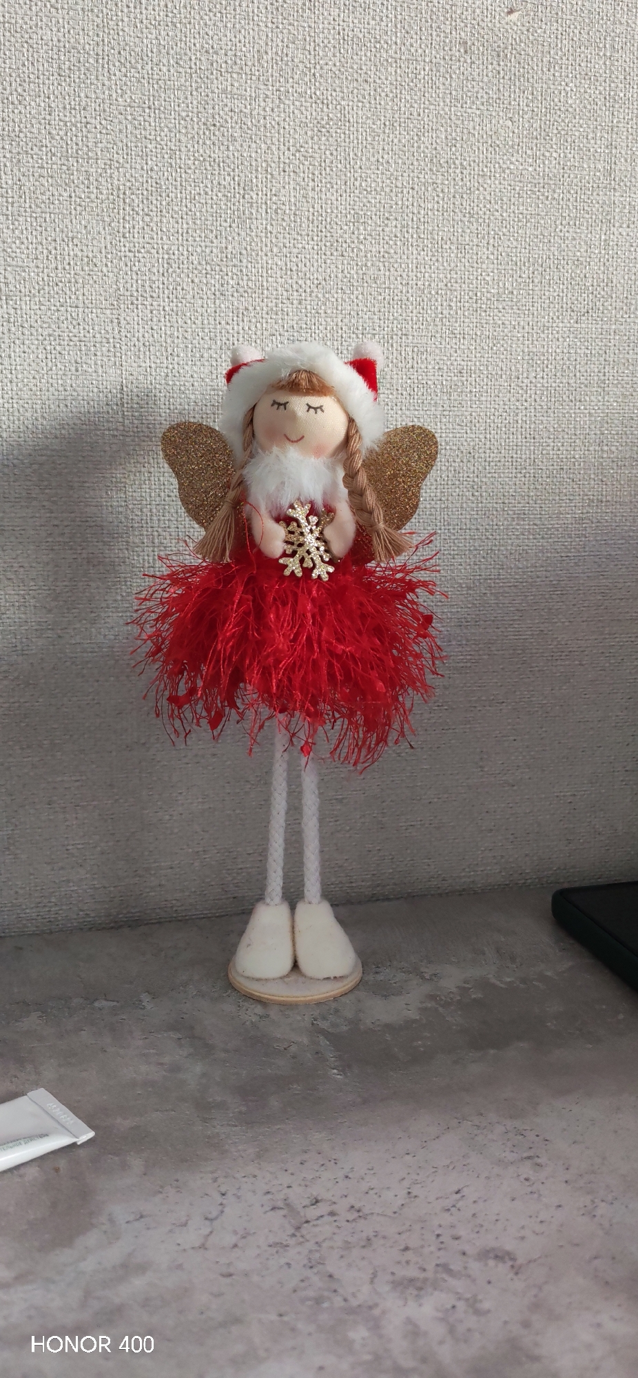 The image shows a decorative angel doll with a red dress and golden wings, standing on a table against a textured wall. The doll is holding a golden snowflake. "HONOR 400" is printed in the bottom left corner of the image.
The location details (Noviy, Russia) were not observable in the image and should not be added.