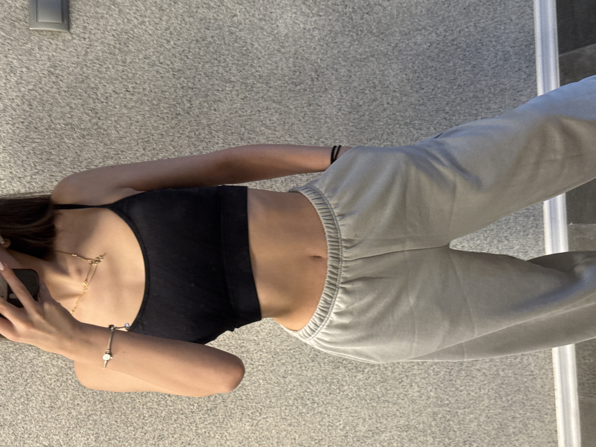 The image, originally rotated ninety degrees counter-clockwise, depicts a person taking a mirror selfie indoors. The subject, seen from the mid-chest to the upper thighs, wears a black ribbed athletic top or crop top and light grey, loose-fitting sweatpants with an elasticated waistband. A golden layered necklace hangs around the person's neck, and a silver-toned charm bracelet is visible on their left wrist. Two black hair ties are worn on the right upper arm. The person's left hand holds a smartphone, partially obscuring their face, as they capture the reflection. The setting appears to be a room with a textured grey carpeted floor. In the background, a grey wall features a light switch in the upper left corner. A white vertical object, possibly a door frame or the edge of a mirror, is visible along the right side of the frame. The midriff is exposed between the top and sweatpants. No specific time of day or weather conditions are discernible from the indoor setting, and no visible text is present in the image. The user provides the context that the location is Sofia, Bulgaria.