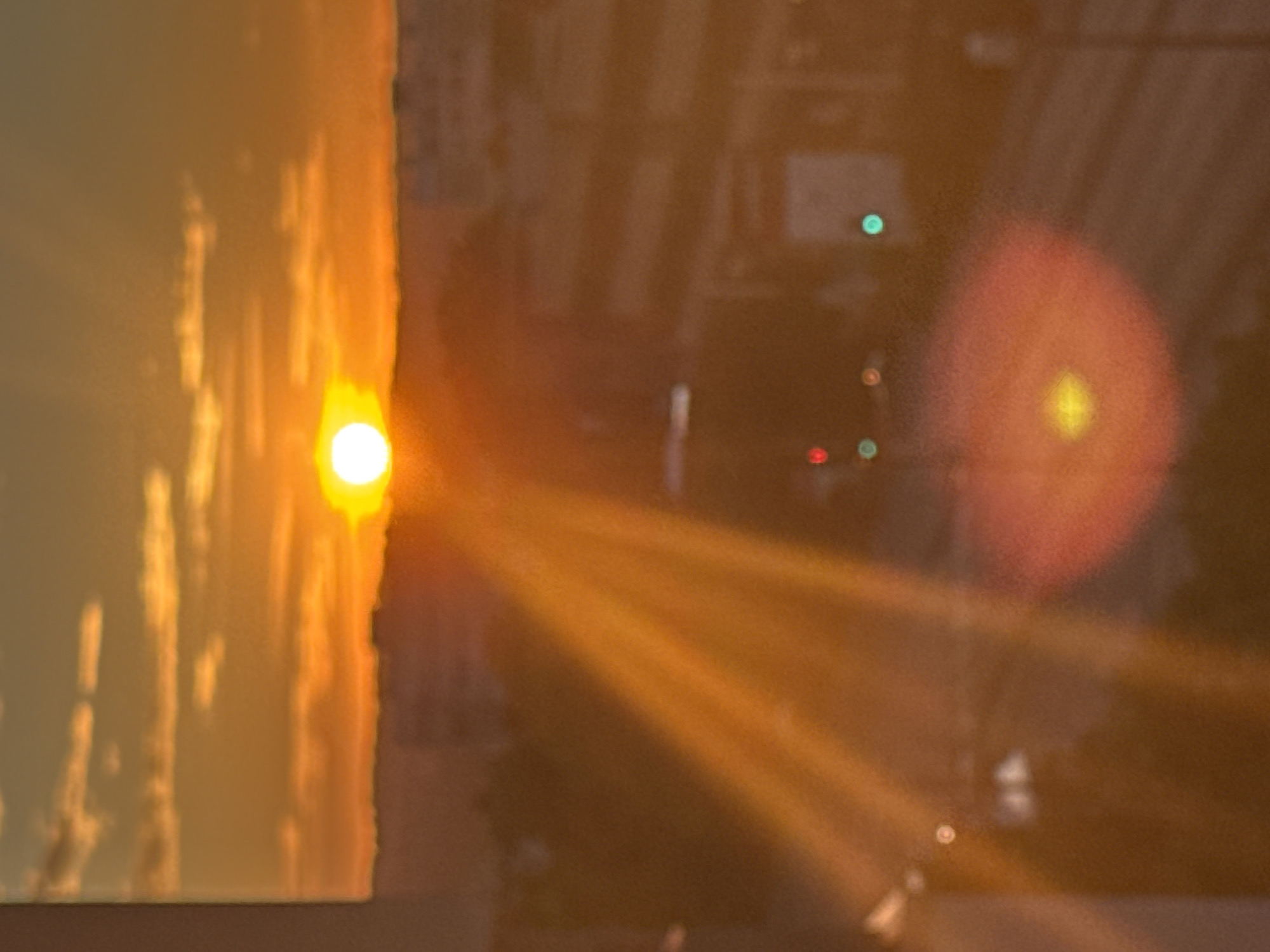 The image depicts a sunset over an urban landscape. The sun, a bright white orb, is low on the horizon, casting warm orange and yellow light across the sky and clouds. Sun rays emanate from the sun, creating a lens flare effect that streaks across the lower half of the image. In the midground, a blurred cityscape is visible, with buildings and what appears to be a road. Several colored lights, possibly traffic signals or building lights, are scattered throughout the scene, with a prominent red and yellow bokeh effect in the foreground. The overall impression is one of a hazy, atmospheric sunset in a city.