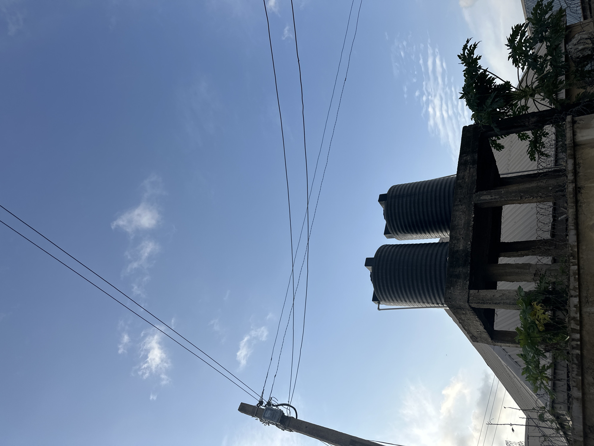 The image depicts a scene on Rukuba Road, Nigeria, featuring a concrete water tank structure supporting two large black water tanks. The structure has visible concrete pillars and a flat roof. In the foreground, a concrete wall topped with barbed wire separates the viewer from a building with a corrugated metal roof and some greenery, including a papaya tree. A utility pole with electrical equipment and wires is visible on the left, with multiple wires stretching across the blue sky, which has scattered white clouds. The overall environment suggests an urban or semi-urban setting in Nigeria.