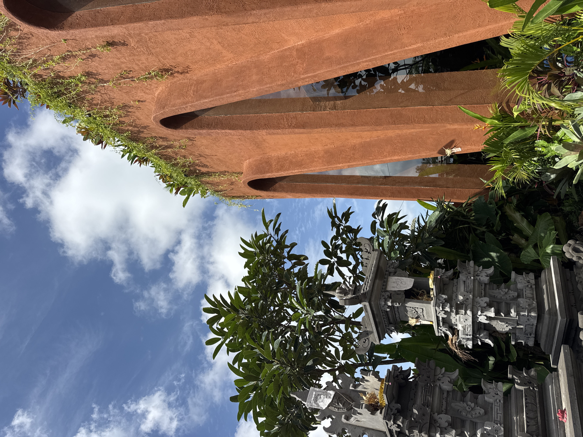 The image shows a close-up, low-angle view of a terra cotta-colored building with arched windows, suggesting it is in a tropical location. Lush green foliage and a large tree with dark green leaves are in the foreground. To the left, two intricately carved stone altars are visible, adorned with offerings. The sky above is a vibrant blue with scattered white clouds. The overall impression is of a tropical setting with architectural elements and religious or cultural artifacts. Given the context of Mengwi, Indonesia, this scene likely depicts a temple complex or a culturally significant site in the region.
