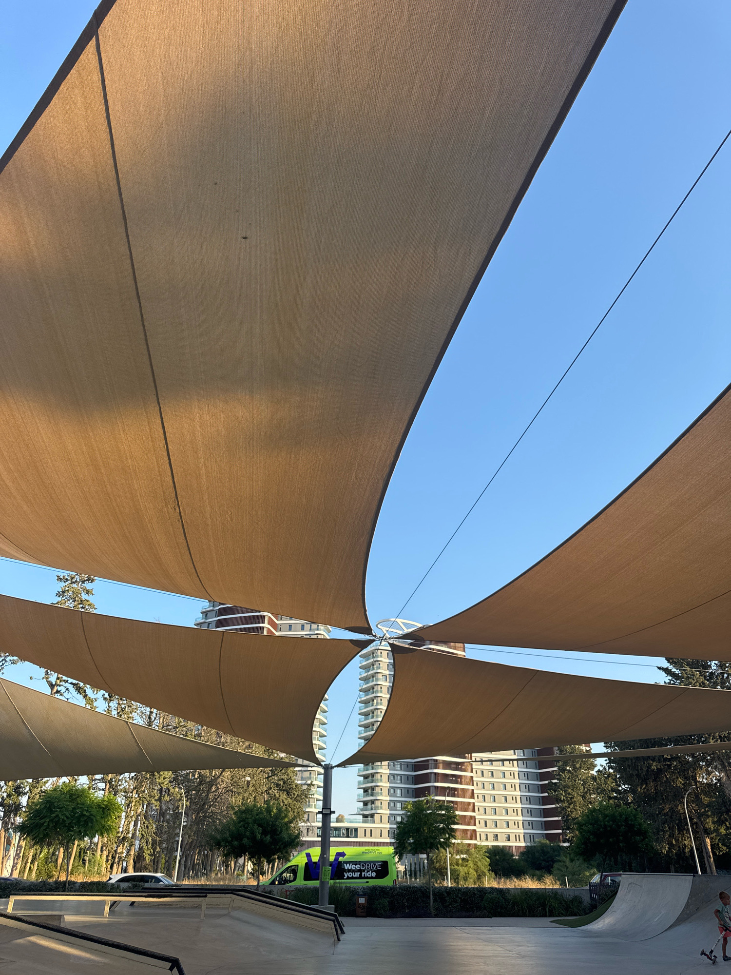 An outdoor perspective shot captures a skate park in Germasogeia Municipality, Cyprus. The scene is dominated by large, beige fabric shade sails suspended in a network above, creating a patterned canopy against a clear blue sky. In the mid-ground, two modern, multi-story buildings with balconies are visible behind a line of trees. A bright green vehicle with text that includes "WeeDRIVE your ride" is parked near the trees. The foreground features the concrete ramps and edges of the skate park. A young boy in shorts and a t-shirt is partially visible on the right side of the skate park, appearing to be in motion or standing on a ramp. A parked car is visible on the left side of the skate park. The lighting suggests it is daytime, possibly late afternoon, given the angle of the shadows.