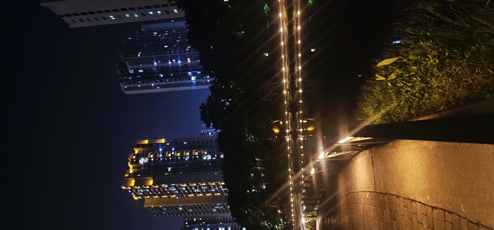 At night in Bishan, China, a paved walkway curves to the left alongside a dark body of water. A wooden railing with illuminated vertical posts separates the walkway from the water. The water reflects scattered lights, and a line of small, lit orbs stretches along the far bank, where trees are silhouetted against the dark sky. Behind the trees, tall buildings with numerous illuminated windows rise into the night. One building on the left is highlighted with golden lights along its upper edges, while the buildings on the right have a cooler, blue-tinted illumination. The sky is dark blue, with no visible celestial bodies. The ground of the walkway appears to be made of reddish brick, with patches of concrete. Grass and some vegetation are visible along the edge of the water. Two small figures, possibly people, are faintly visible in the distance along the lit path on the far side of the water.