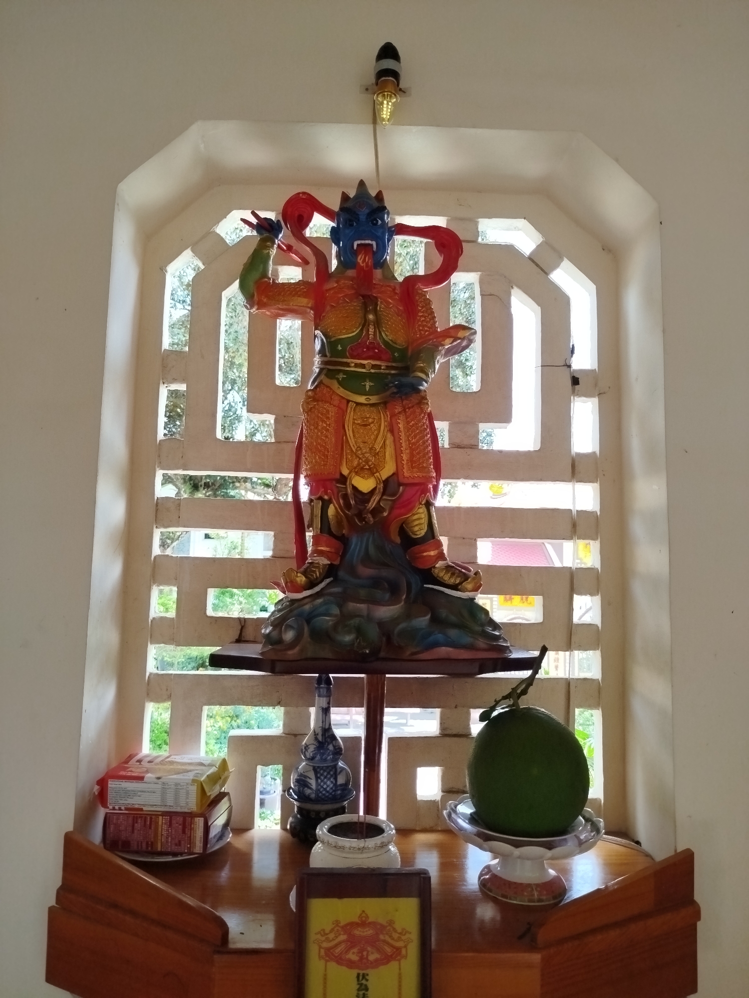 The image shows a small altar with a statue of a deity. The statue is blue-skinned with red accents and wears ornate armor. It stands on a sculpted base depicting clouds. Above the statue, a light bulb hangs from the ceiling. The altar is made of wood and holds various items, including a stack of packaged goods, a blue and white vase, an incense burner, a framed object with writing, and a large green fruit on a pedestal. The altar is positioned in front of a window with a decorative grid pattern, revealing a view of trees and buildings outside. The setting is in Cam Ly - Đà Lạt, Vietnam.