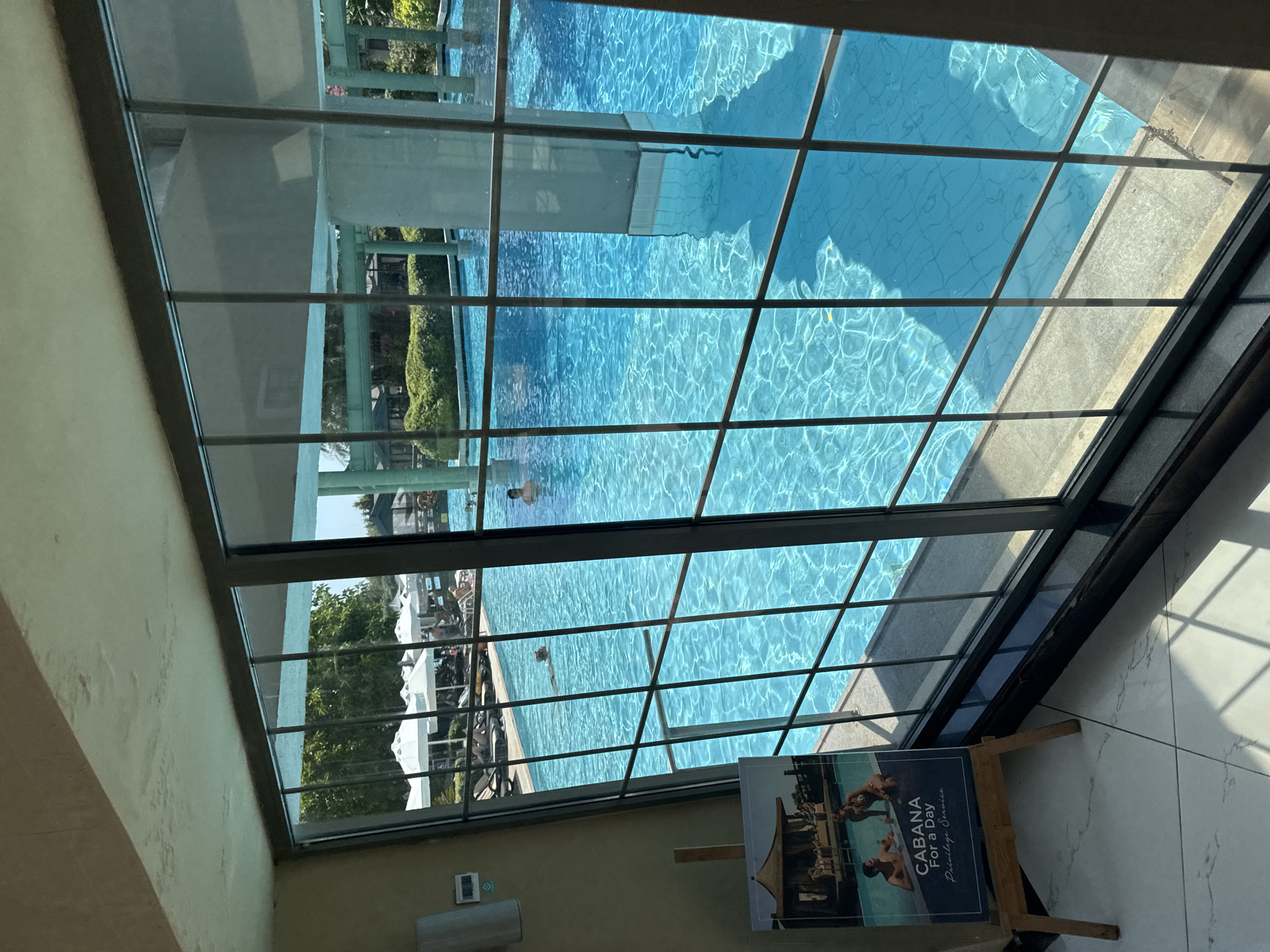 This is a photograph taken indoors, looking out through a large, gridded window at a swimming pool area at the Titanic Beach Lara hotel in Aksu, Turkey. The pool is large and filled with bright blue water, with sunlight reflecting off the surface. Several people are visible in the pool, swimming or relaxing. Outside the window, there are white umbrellas shading lounge chairs. The interior of the room features light-colored walls and flooring with a subtle marble pattern. A wooden easel stands in the foreground, displaying a brochure advertising "CABANA For a Day Privilege Service" with an image of a couple by a pool. The window frame is dark, and reflections from the interior are visible on the glass. The overall impression is one of a luxurious resort setting on a sunny day.