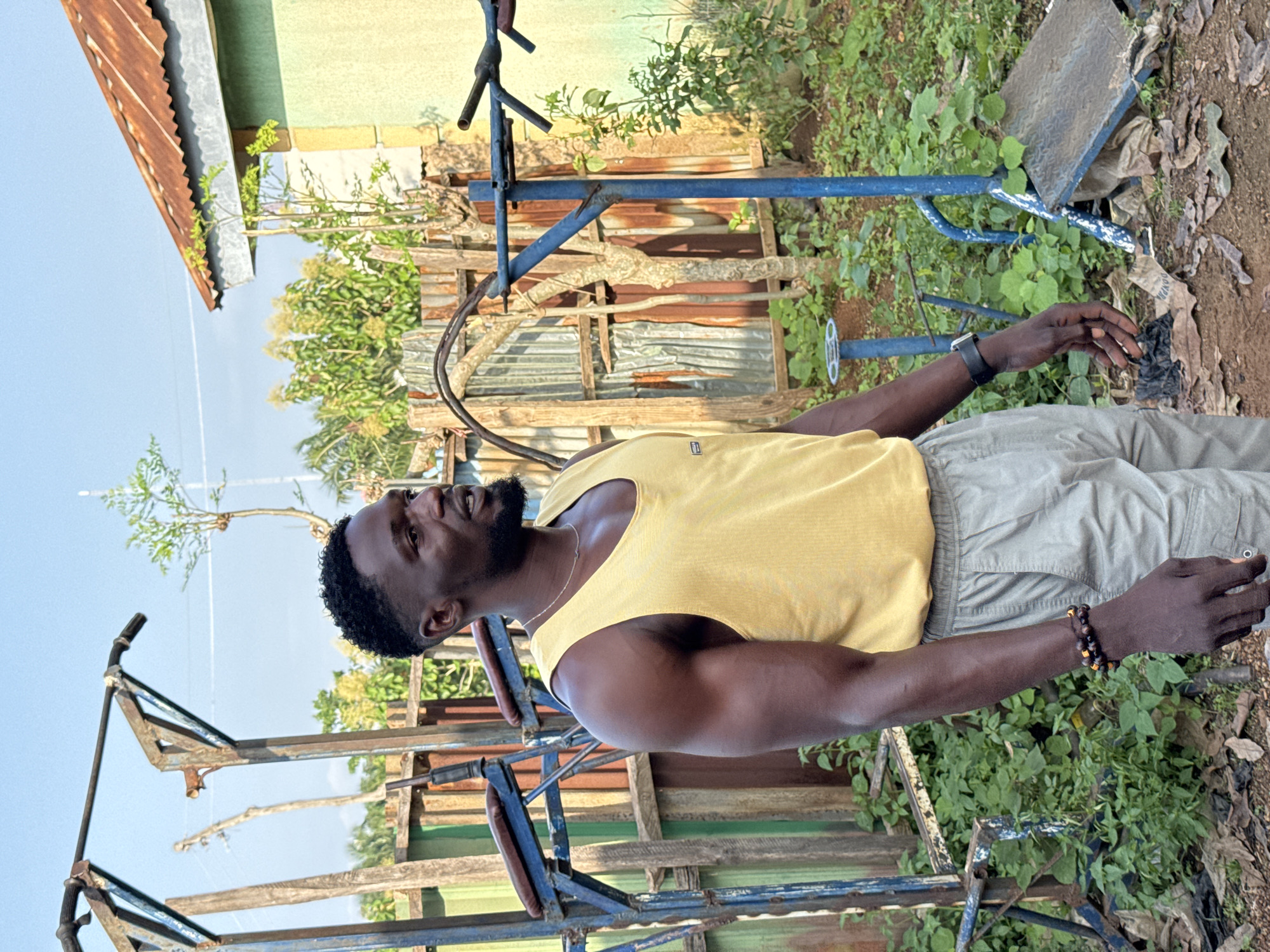 A Black man with a short, dark beard and a fade haircut stands outside in daylight, looking to his left. He wears a yellow sleeveless shirt and light gray cargo shorts. On his left wrist, he has a black smartwatch, and on his right wrist, two beaded bracelets. In the background, a rusted metal structure, possibly exercise equipment, stands amidst overgrown green vegetation and some corrugated metal sheeting. A green building with a corrugated, rusty roof is visible to the right. The ground is covered with dirt, leaves, and some debris. The sky above is clear and blue. This scene appears to be in a somewhat dilapidated outdoor gym area in Akwanga, Nigeria.