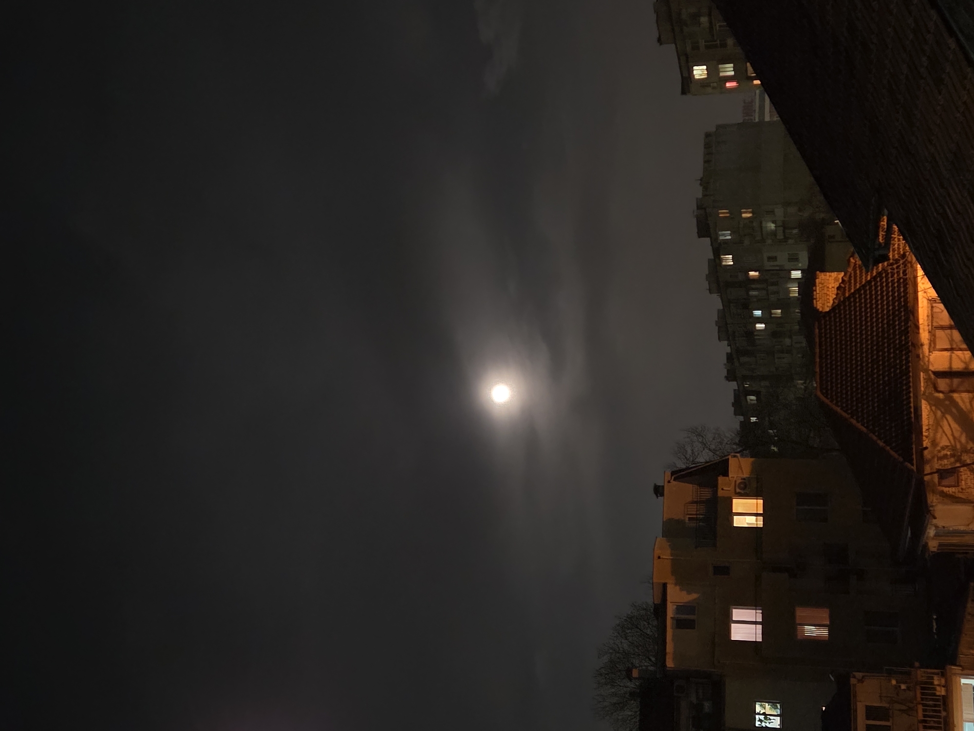 An outdoor night scene, oriented vertically, features a brightly illuminated moon centered in the sky. Light cloud cover surrounds the moon. To the right, multiple residential buildings of varying height are visible, including what appears to be the roof of a house closest to the viewer. Several windows in the buildings are illuminated. Bare trees are interspersed between the buildings. The image appears to be taken from a low vantage point, perhaps at street level. Given the residential architecture, the location is likely Varna, Bulgaria.