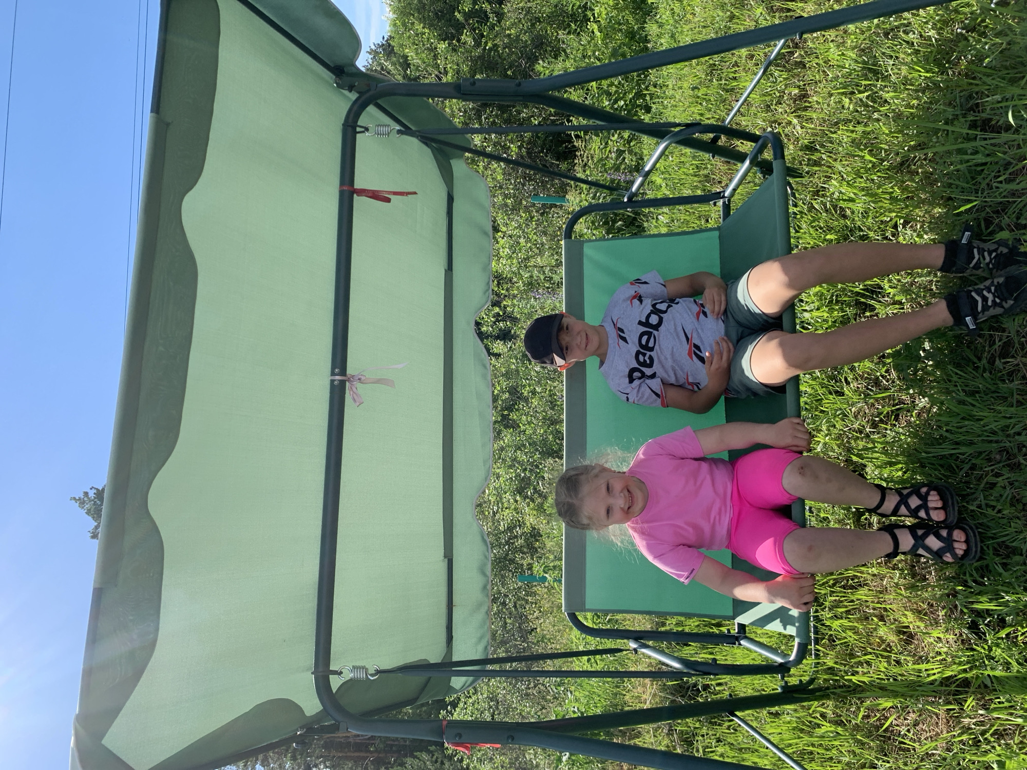In an outdoor scene, two children are sitting on a green swing set. A boy, wearing a black baseball cap and a grey t-shirt with "Reebok" printed on it in black and red, is seated on the right side of the swing. He has dark hair and is smiling. Next to him on the left, a young girl with blonde hair is wearing a pink t-shirt and bright pink shorts. She has a wide smile and is looking towards the camera. Both children are wearing sandals. The swing is under a light green canopy, and the background consists of lush green grass and trees, suggesting a sunny day. The image appears to be taken in a residential backyard or a park. The overall mood is cheerful and relaxed.