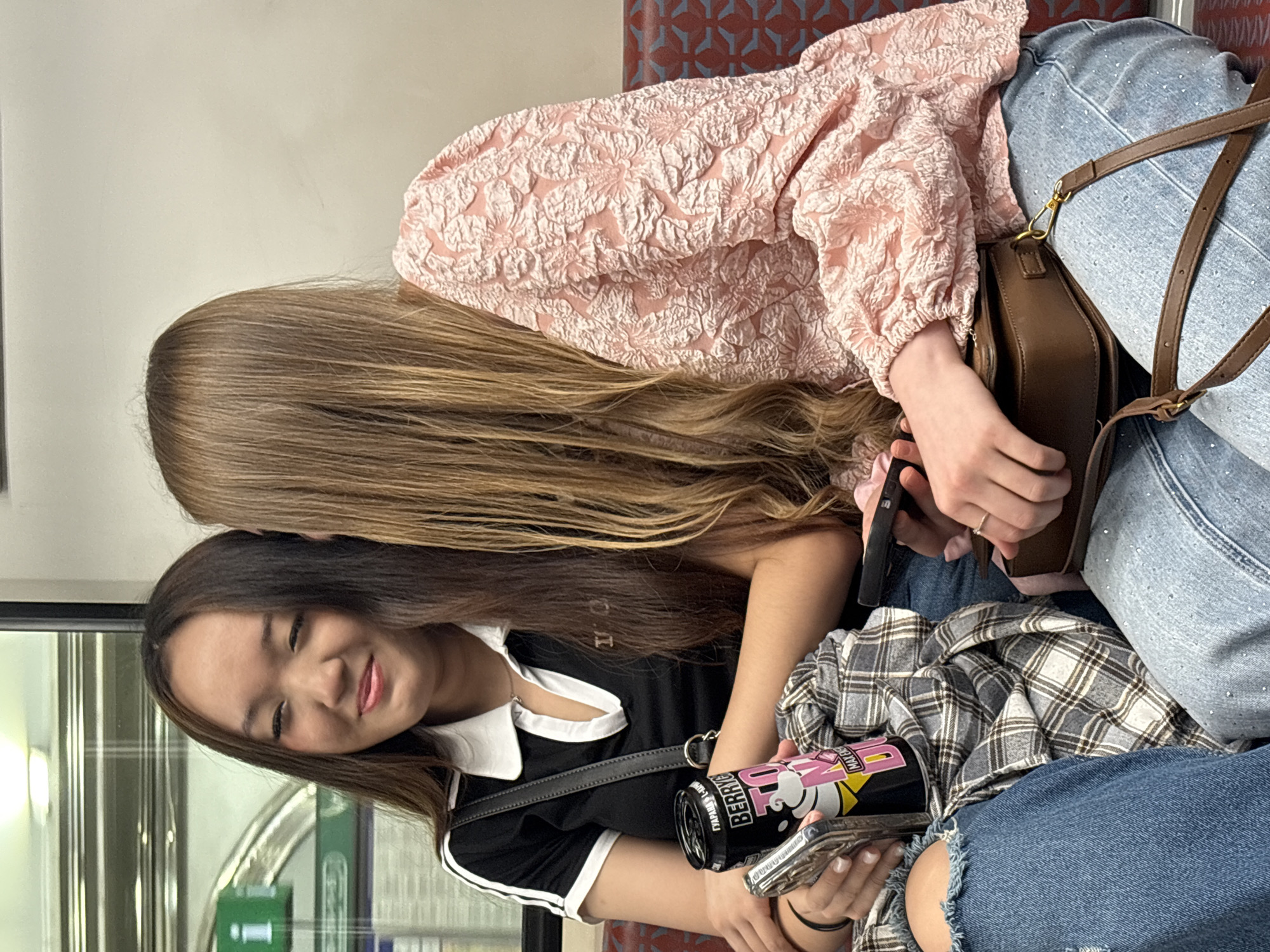 Two young adult females are seated closely together on what appears to be public transit within the city of Saint Petersburg, Russia, indicated by Cyrillic text on an object and the environment consistent with a metro or train car.
The woman on the left has long, dark brown hair with lighter brown highlights, styled straight with a center part. Her skin tone is consistent with East or Southeast Asian ethnicity. She wears subtle eye makeup and has a slight, closed-lip smile, looking towards her left. Her attire consists of a black short-sleeved polo shirt with a white collar and white stripes on the sleeves. A grey and white plaid shirt is draped over her lap, revealing distressed blue denim jeans on her right leg. She holds a black beverage can labeled with "BERRY" and "MAX ENE," featuring Cyrillic text "ГУАРАНА" (Guarana) and "ЯГОДА" (Berry). Simultaneously, she holds a silver-colored smartphone. A black shoulder bag strap crosses her right shoulder. Her left arm rests across the adjacent woman's arm/lap.
The woman on the right has long, straight, light brown hair that predominantly obscures her face from the viewer's perspective. She appears to be looking downwards or towards her right. She wears a light pink long-sleeved blouse with a prominent textured, embossed floral or botanical pattern. Her lower garments are light blue denim jeans embellished with small, reflective elements (rhinestones) on the upper thigh. She holds a dark-colored smartphone in her left hand and wears a thin silver ring on her left ring finger. A small, dark brown leather crossbody bag with a gold-toned clasp and buckle rests on her lap.
The seating features upholstered red and blue geometric patterned fabric visible on the right. The background on the right is a plain, light beige wall. On the left, a window shows a portion of a green "i" (information) sign, indicative of a station platform, and reflections of the vehicle's interior. Overhead, linear fluorescent lighting illuminates the scene. The women are in close proximity, suggesting a personal interaction.