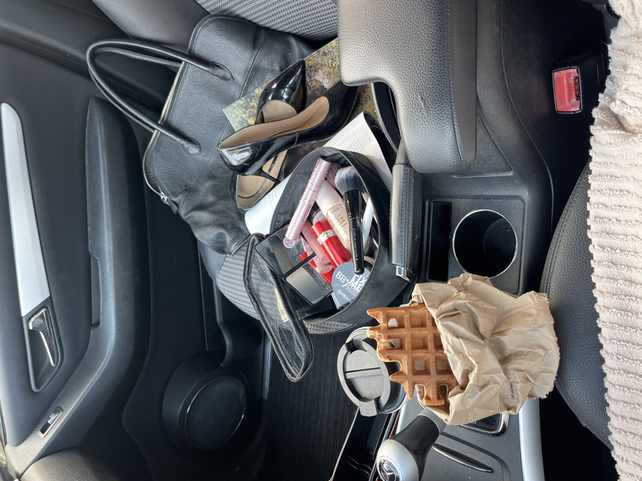 The image displays the interior of a vehicle, specifically focusing on the front passenger seat and the adjacent center console. The vehicle's interior is predominantly black and dark grey. The passenger seat is upholstered in a grey patterned fabric. A Mercedes-Benz logo is discernable on the automatic gear shift knob.
On the passenger seat, a black leather handbag with two handles is positioned, along with a pair of black high-heeled shoes, which appear to be patent leather with ankle straps, resting on a folded piece of paper or magazine. Adjacent to these items is an open, black zippered makeup bag containing assorted cosmetic products: multiple tubes of lipstick/lip gloss (red, pink, and light pink shades), a pink-cased mascara, foundation/BB cream bottles, various makeup brushes, and a compact with a mirror.
On the center console, a light brown, rectangular waffle, exhibiting a distinct grid pattern and an irregular top edge, is held upright within a crumpled light brown paper bag. This assembly is situated in one of the vehicle's cupholders. Other visible elements of the center console include an automatic gear shift knob, a manual handbrake lever with a chrome button, additional cupholder openings, and a red-buttoned seatbelt buckle receiver. No individuals are present in the scene. This environment is located within a vehicle in the city of Budva, Montenegro.