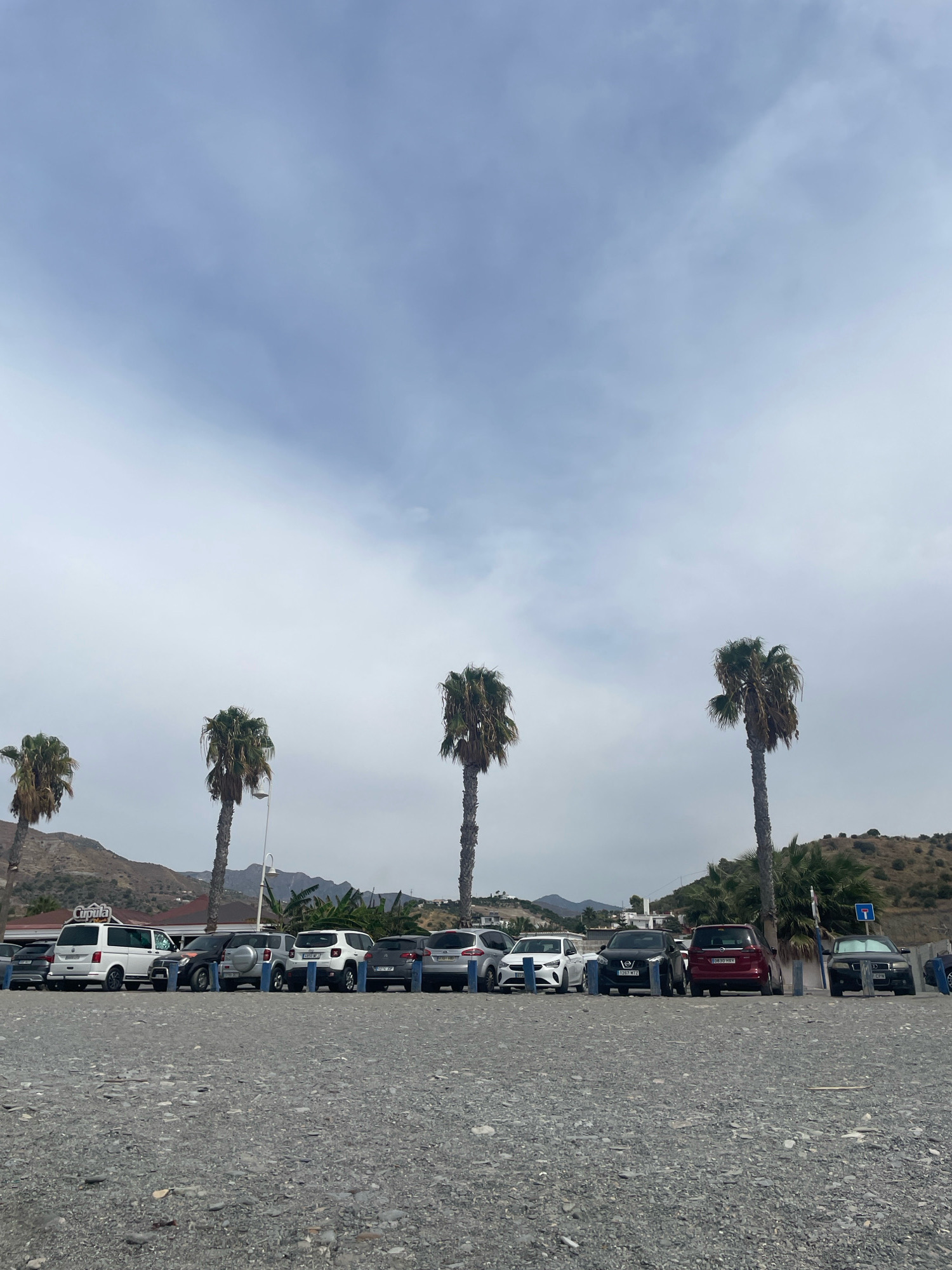 In the city of La Herradura, Spain, a gravelly parking lot is situated in front of a row of parked cars, with palm trees and hills in the background under a cloudy sky. The vehicles are parked in a line from left to right: a silver SUV, a white van with "CUPULA" written on the side, a black SUV, a white SUV, a dark gray SUV, a white hatchback, a black sedan, a dark red minivan, a black sedan, and a silver sedan. A blue traffic sign with a white line and a white arrow pointing right is visible next to the dark red minivan. The ground is covered in small, dark gray pebbles. The sky is overcast with soft, white clouds. The background features rolling hills with sparse vegetation and some buildings at the base. Several palm trees are scattered throughout the scene, with their fronds appearing weathered. There are no people visible in the immediate foreground of the image, but a person is partially visible in the far right, near the silver sedan.