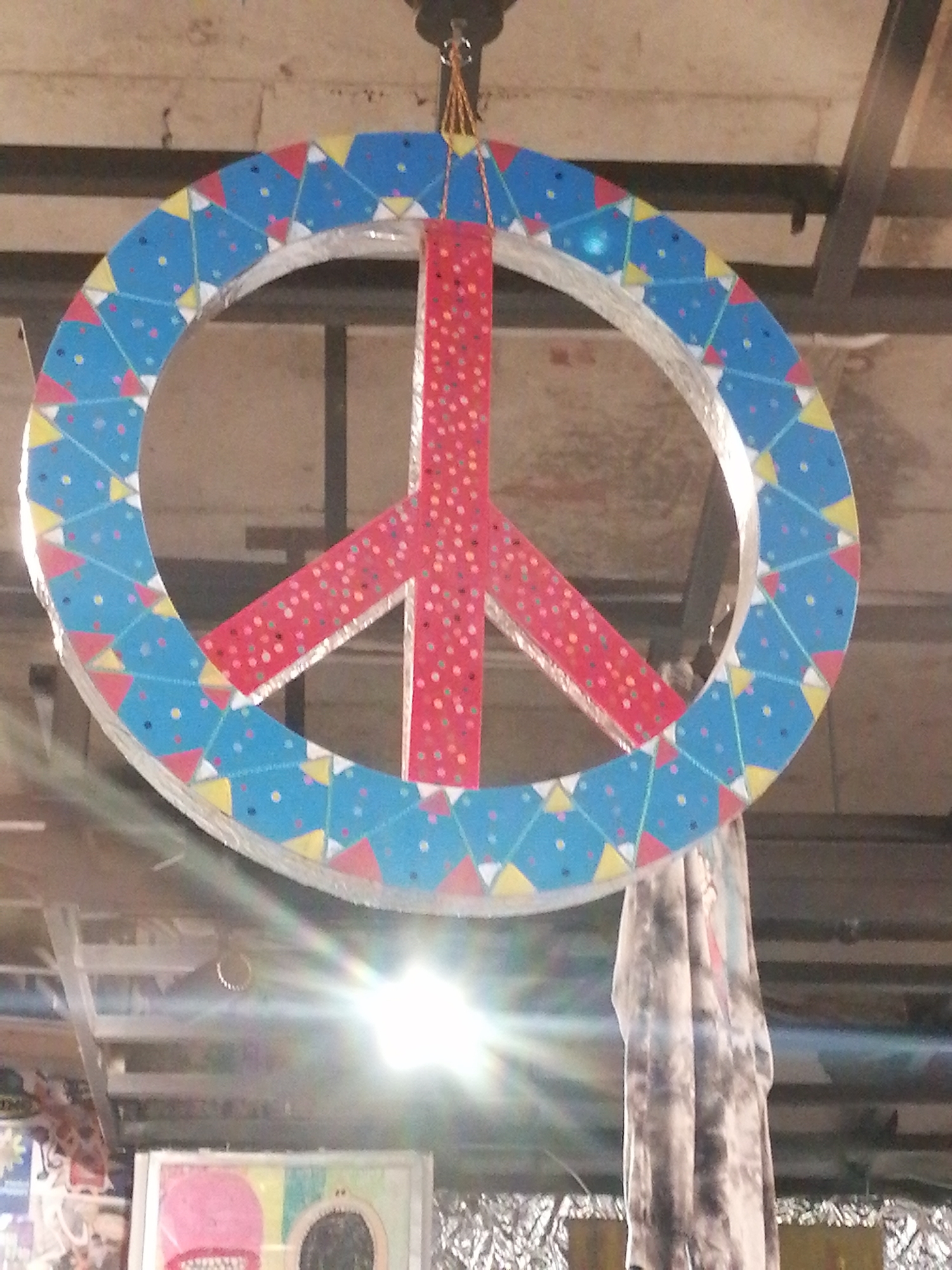 The image shows a large, decorated peace sign hanging indoors. The peace sign is circular with a red symbol inside. The outer ring of the peace sign is decorated with blue sections containing small, colorful dots and triangular patterns in yellow, red, and white. The red symbol inside the peace sign is also decorated with small, colorful dots. The peace sign is suspended by a thin cord from a ceiling structure made of metal beams. There is a bright light source creating a lens flare in the lower part of the image, obscuring some details of the environment below. In the background, a piece of fabric with a tie-dye pattern hangs down, and some indistinct objects are visible. The overall impression is that of an indoor space, possibly a workshop or a decorated area, in Da Lat, Vietnam.