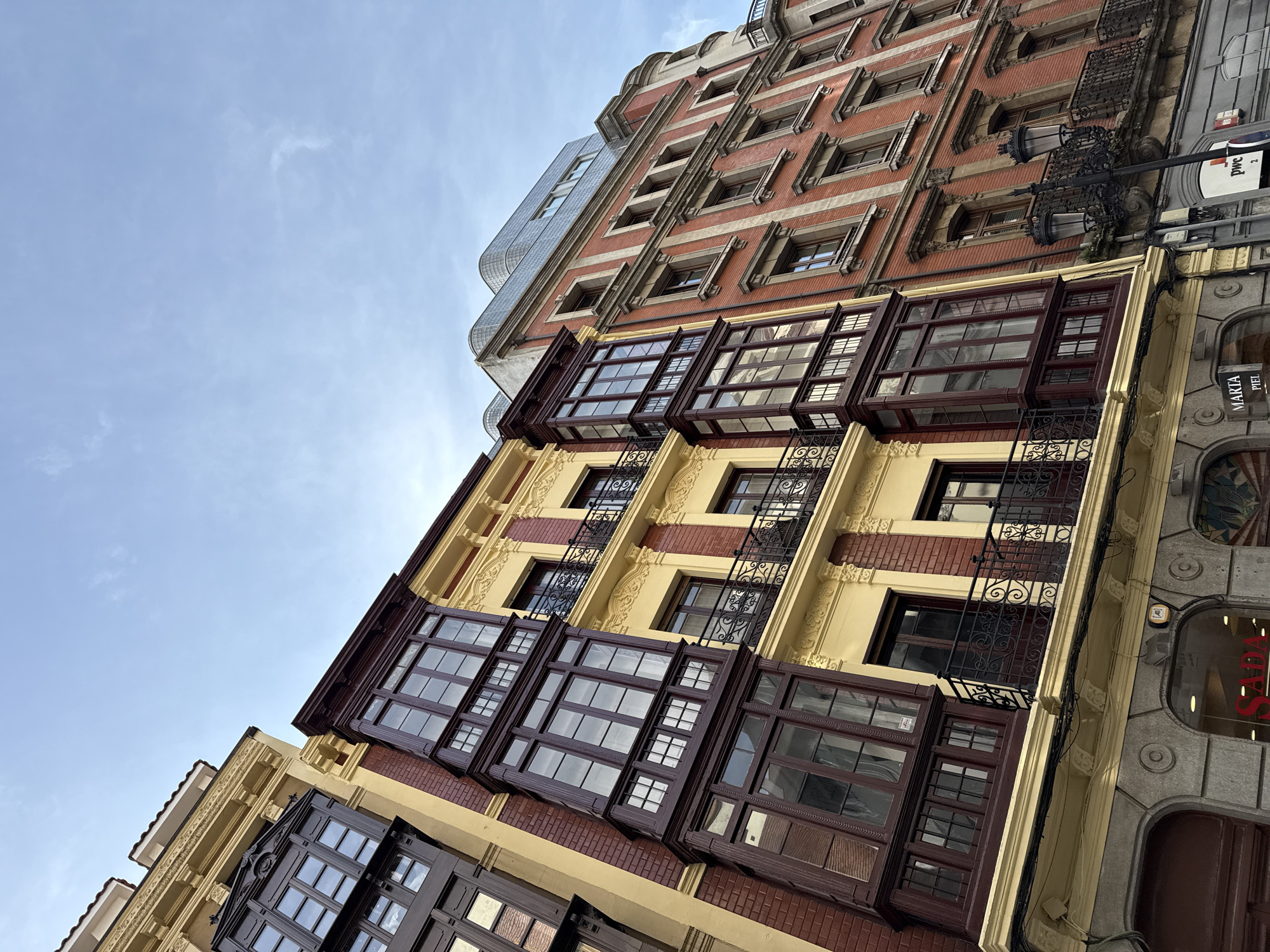 The image shows a low-angle view of several buildings in Oviedo, Spain. The buildings are of varying architectural styles and colors. The leftmost building has a modern, glass facade. The adjacent building is taller, with red brick and decorative stone elements, featuring numerous windows. In front of this brick building is another building, displaying large bay windows with dark brown trim, set against a yellow and red brick facade. Black iron fire escapes are attached to this building. On the right side of the image is a section of a building with a grey stone facade and visible storefront windows. The sky above is light blue with some clouds.
