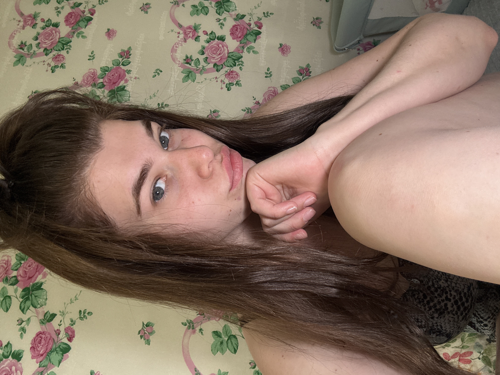 A young woman with long brown hair and blue eyes is lying on a floral patterned surface. She is fair-skinned and has her right arm bent, with her hand near her face. The floral pattern consists of roses and green leaves. The background appears to be a wall or upholstered surface with the same floral pattern. Part of a gray object is visible in the upper right corner. The location of Мещёры, Russia cannot be determined from the image.