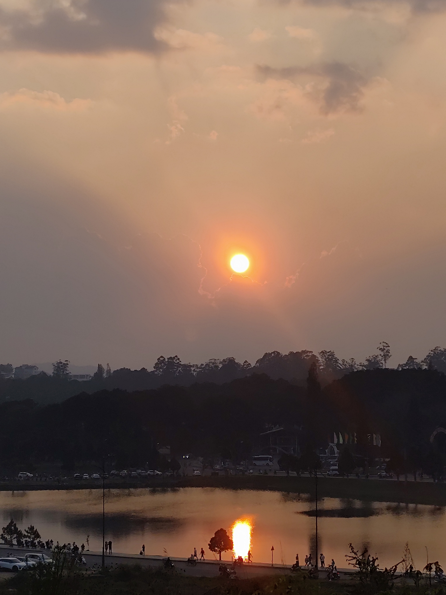 The image depicts a sunset over Xuan Huong Lake in Da Lat, Vietnam. The sky, occupying the upper two-thirds, displays an orange-brown gradient with scattered clouds; a bright, circular orange sun is prominent slightly right of center, casting a visible reflection on the lake's surface. Silhouetted forested hills form the distant horizon line. In the midground, Xuan Huong Lake reflects the bright sun in a vertical orange-yellow column. Along the lake's edge, a paved promenade or road is populated by numerous individuals whose silhouettes are visible walking, standing, and interacting. Several parked and moving vehicles, including cars and motorcycles, are present on the road. A tall streetlamp post stands near the center. Across the lake, a more distant shoreline features additional parked vehicles, including a bus, and structures with multiple flags. Dark foliage partially obscures the immediate foreground.