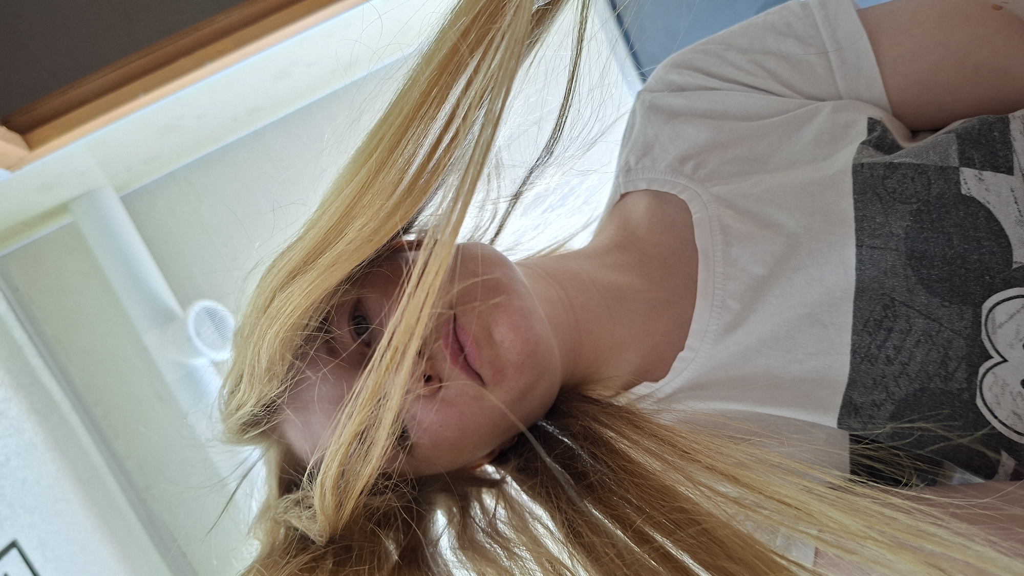 A young person with blonde hair and light skin is shown from a low angle, looking upwards. Their hair is windblown and obscures parts of their face. They are wearing a white t-shirt with a dark graphic print. The background appears to be indoors, with a white ceiling and a window frame visible. There are no discernible features that indicate the location is in the city of Bolshoy Bukor, Russia. The image is an unposed selfie.