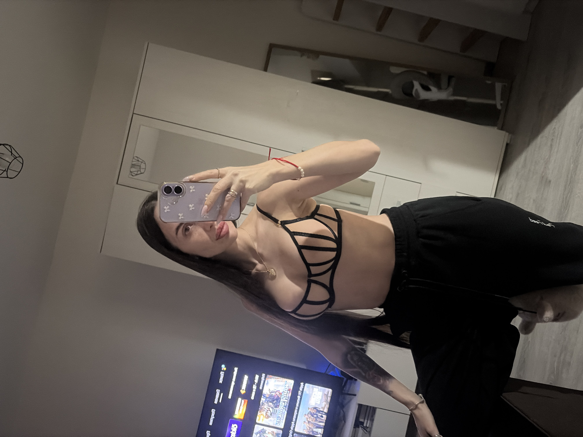 A woman is posing for a selfie in a room with a TV, a wardrobe, and a mirror. She is wearing a black strappy bra and black sweatpants. The TV screen is showing various images. The location is likely indoors. No evidence to indicate this photo was taken in Saint Petersburg, Russia.