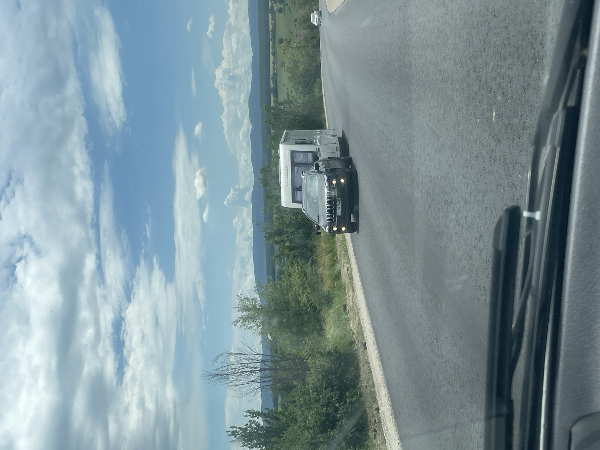 The image shows a view from inside a vehicle on a road near Probuda, Bulgaria. In the distance, there are slightly rolling, tree-covered hills. A black car is overtaking a white caravan on a two-lane asphalt road. The sky is light blue with scattered clouds. The camera is looking out of the windshield from within the vehicle.