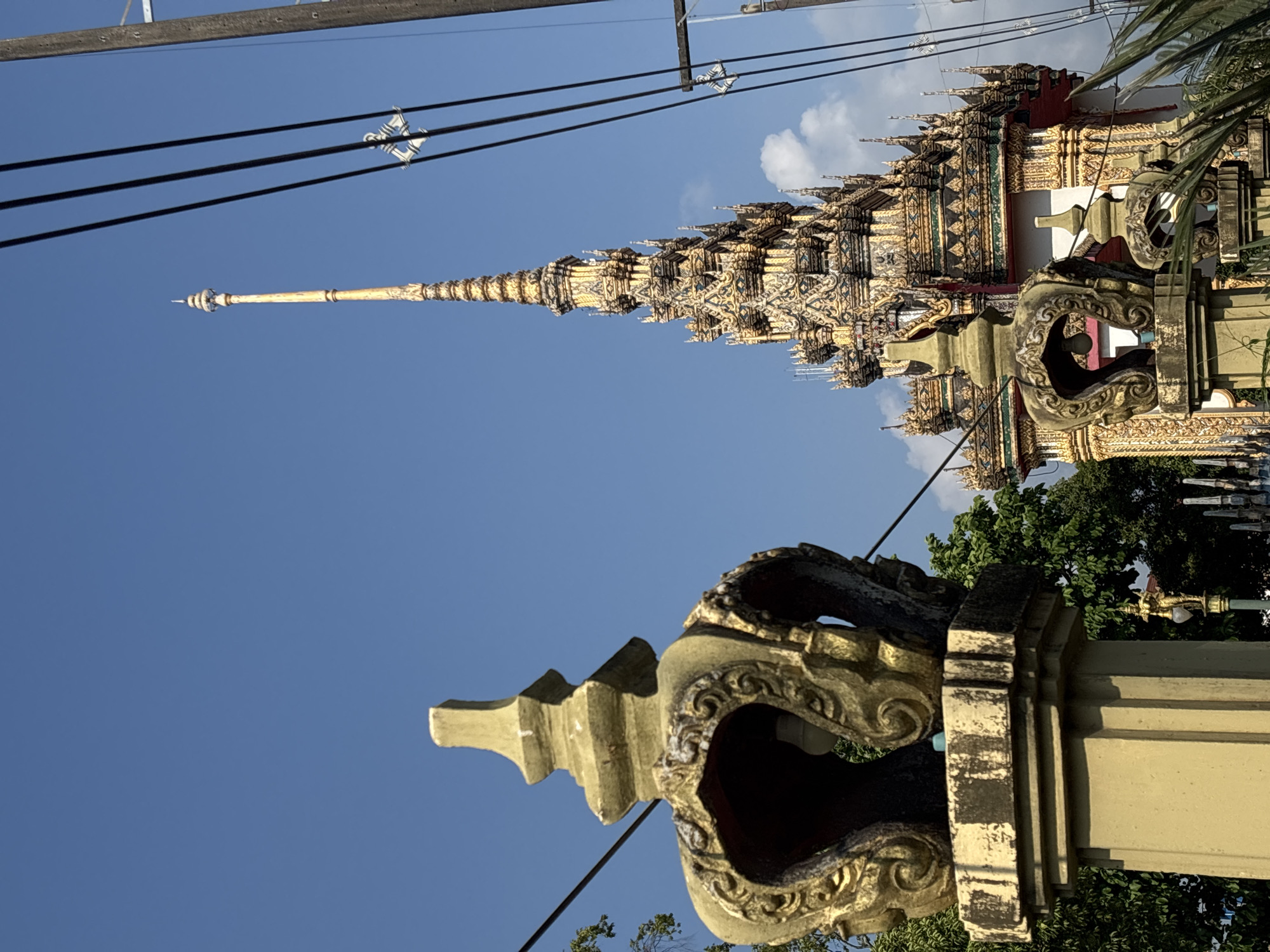 A vibrant, daytime shot captures a tall, ornate golden spire of a temple structure against a clear blue sky, partially oriented horizontally due to the image's rotation. The spire, indicative of Thai architecture, is richly detailed with intricate golden patterns, multiple tiers, and small, spire-like projections. Below the main spire, the upper sections of a larger temple building are visible, featuring similarly elaborate gold and mosaic decorations, along with red and green accents.
In the foreground, an ornate, weathered concrete or stone decorative element, possibly part of a gate or balustrade, occupies the lower portion of the frame. It displays intricate scrollwork and a deep, shadowed opening. Above, several dark power lines with white insulators stretch across the sky, contrasting with the temple's grandeur. Green foliage from trees is visible on the right side and in the background, peeking from behind the foreground structure and temple. The bright sunlight suggests a clear day, likely late morning or afternoon. The architectural style strongly points to a Buddhist temple in Thailand, aligning with the given location context of Choeng Thale. There are no people or visible text.