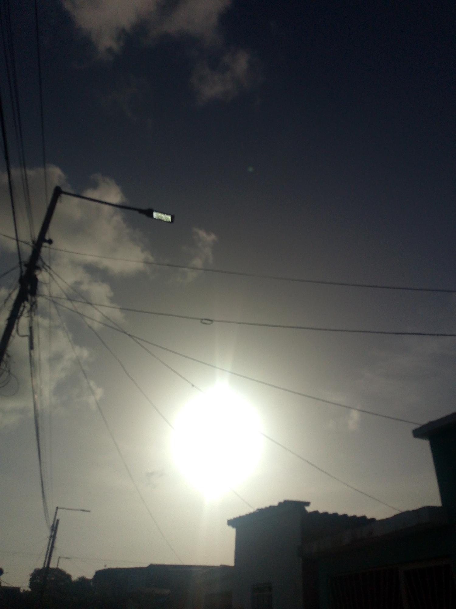 A low-angle, blurry photograph taken in Juan Griego, Venezuela shows a street light fixture on a dark pole adorned with multiple electrical wires against a cloudy sky with the sun shining brightly behind the clouds. The sun's glare is so intense that it appears as a white orb in the center of the frame, obscuring details of the sky and any potential objects behind it. In the foreground, silhouetted against the bright sky, are the dark outlines of rooftops and buildings, suggesting an urban or semi-urban environment. The overall impression is of a hazy, overcast day with a strong light source.