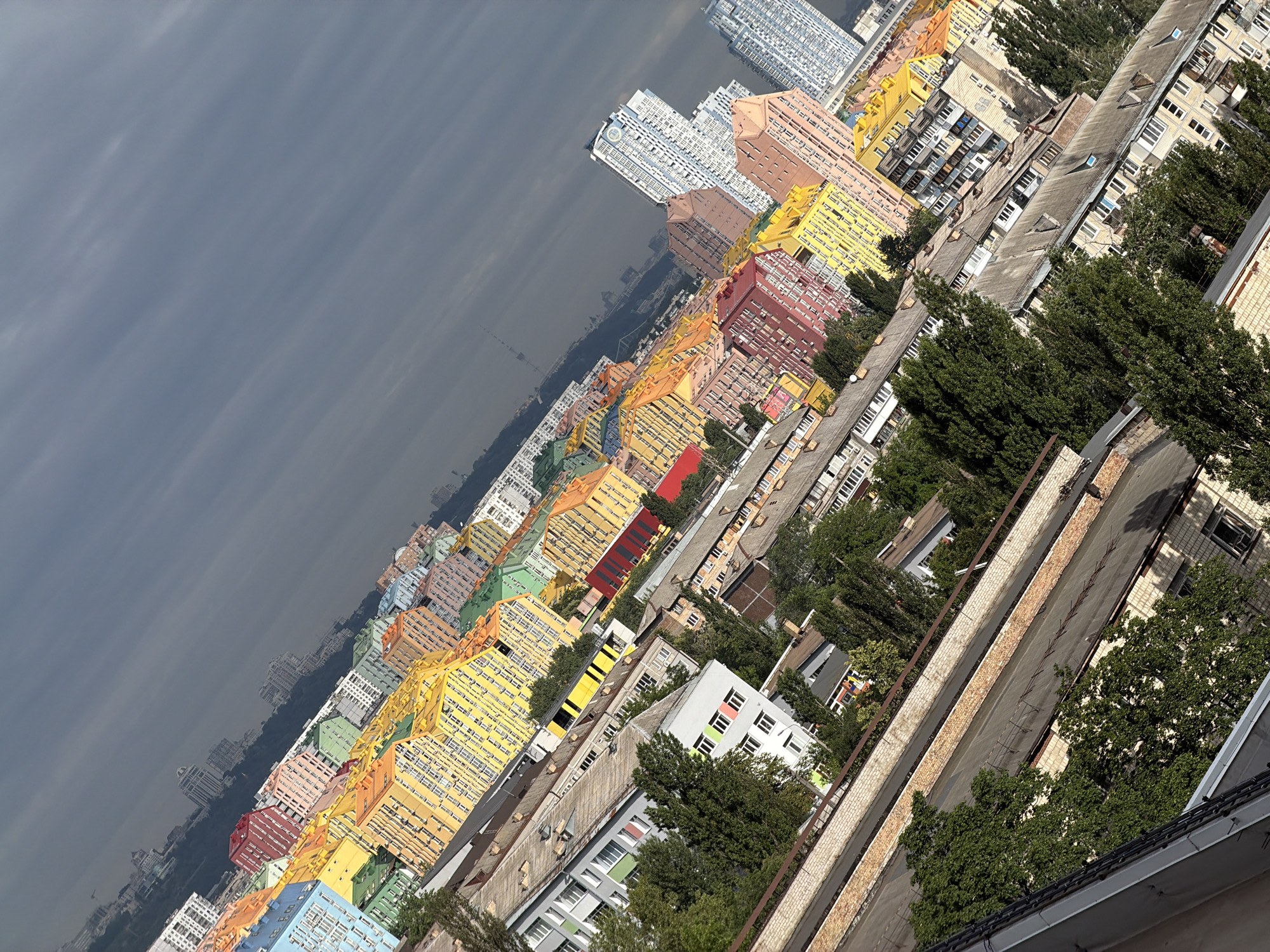 The image captures a panoramic view of the colorful residential buildings in the "Comfort Town" complex in Kyiv, Ukraine. The buildings are painted in vibrant hues of yellow, orange, pink, green, and blue, creating a striking visual contrast against the overcast sky. The perspective is tilted, giving a dynamic, almost abstract feel to the scene. In the foreground, older, more muted buildings and trees suggest a layering of urban development. Further in the distance, taller, modern apartment blocks, some white and blue, dominate the skyline, indicating the ongoing growth and diversification of the city. The scene appears to be taken from a high vantage point, looking down and across the urban landscape. There are no people or vehicles visible in the image.
