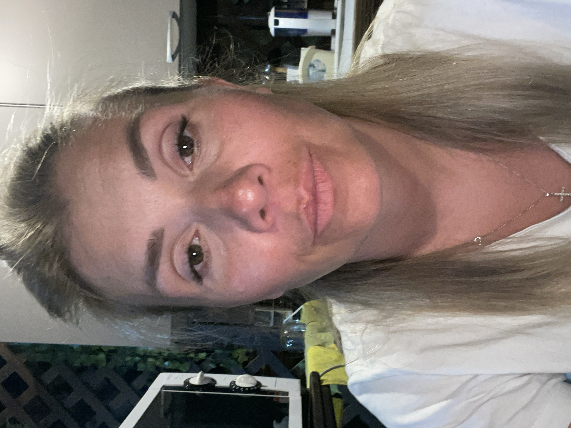 A medium close-up, indoor selfie shows a fair-skinned woman with blonde hair pulled back. She is wearing a white t-shirt and a silver cross necklace. Her face is illuminated from the front, with no shadows visible. The background is out of focus, but appears to be a kitchen or dining area. To the left, a white microwave is visible. To the right, a blue and white dispenser is on a shelf. A dark green lattice structure with some foliage is visible behind the microwave. The lighting is warm and natural.