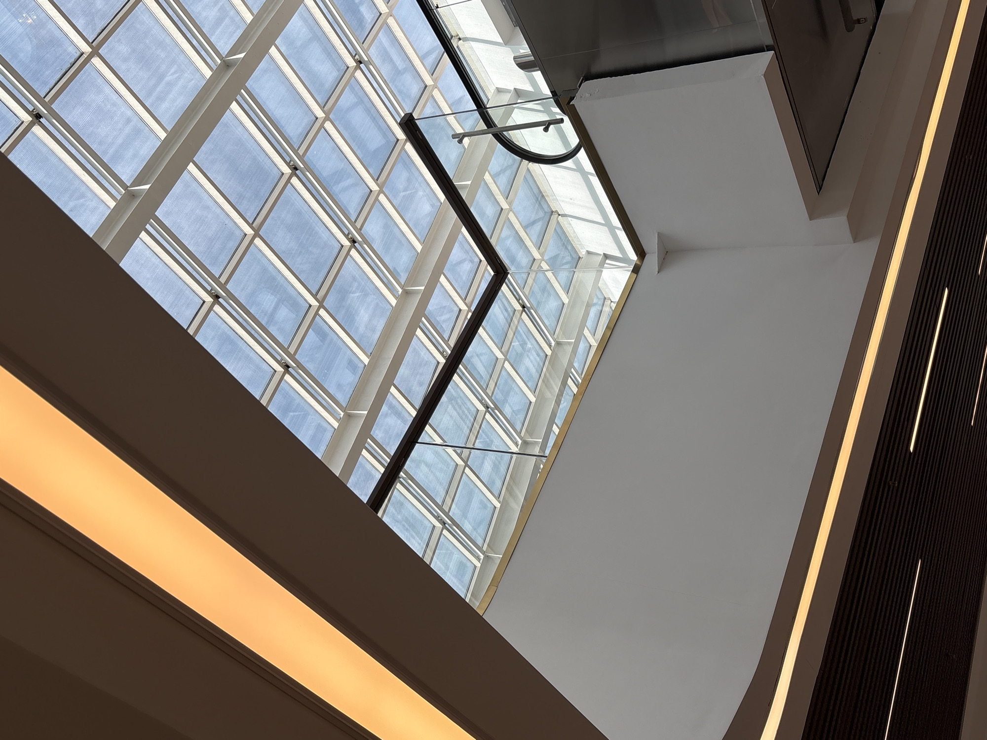 The image shows a modern interior with a glass ceiling and contemporary lighting elements. The ceiling is a grid of square glass panels supported by a white frame, allowing natural light to filter through. Below the glass ceiling, there is a white wall and an escalator with a glass railing. Warm, yellow light emanates from long linear fixtures along the ceiling and the upper edge of the wall. The lower part of the image reveals a darker, ribbed surface with more linear lighting. The scene depicts an indoor public space, possibly a shopping mall or an airport terminal, characterized by clean lines and bright illumination. The image does not contain any people or specific identifiable landmarks that would suggest a location in Saint Petersburg, Russia.