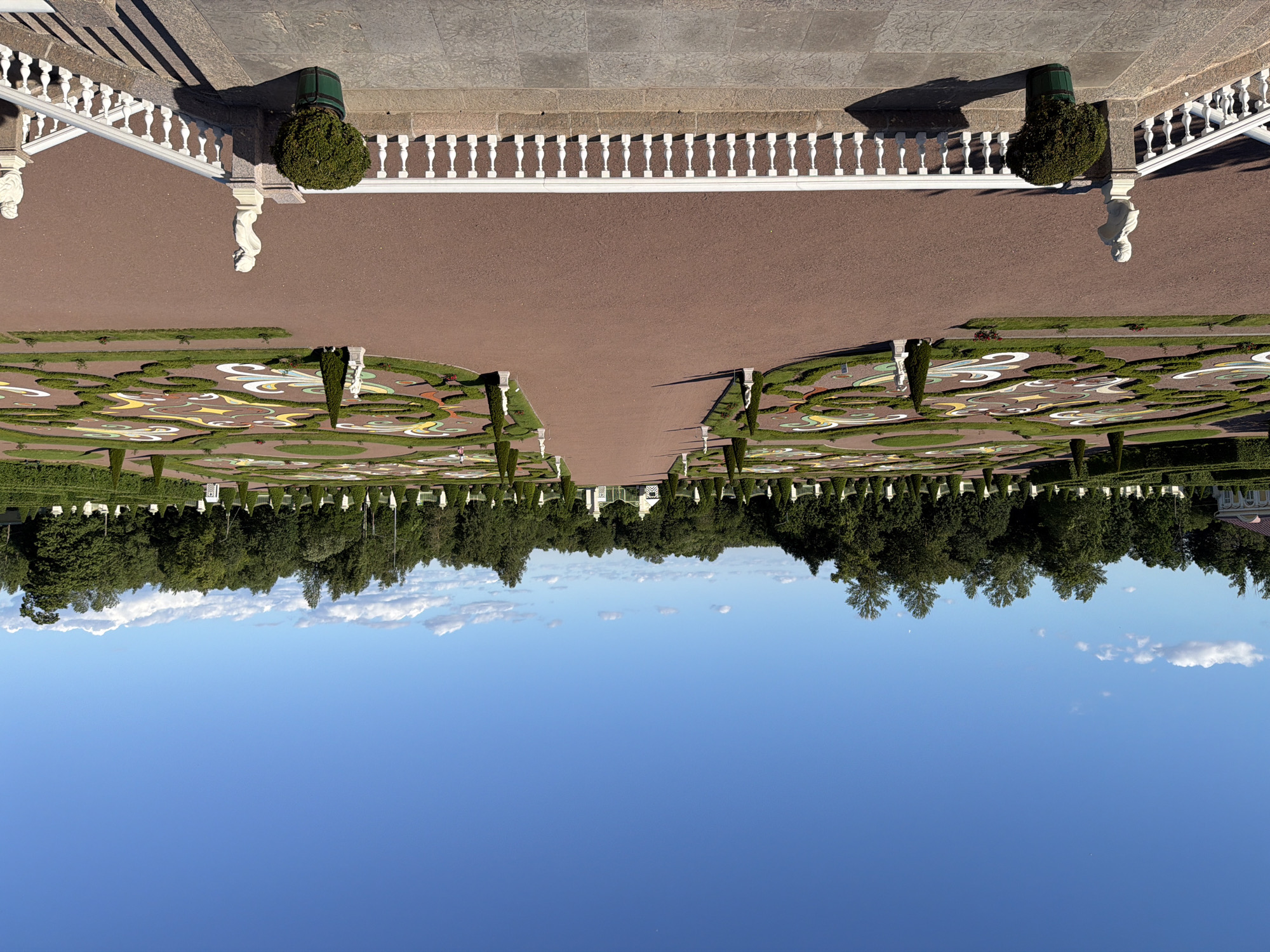 The image depicts a meticulously manicured formal garden, characterized by intricate parterre designs formed by low green hedges. These geometric patterns are filled with contrasting colored gravel and mulches, creating a visually striking mosaic. The garden is symmetrical, with a central pathway leading towards a distant building, flanked by rows of manicured trees and ornate white balustrades. Sculptural elements, including busts and topiaries in planters, are strategically placed throughout the garden, adding to its classical aesthetic.
In the foreground, there is a broad expanse of clear blue sky with a few scattered white clouds. The perspective of the image is unusual, suggesting it was taken from a high vantage point and then rotated, or perhaps it is a reflection. The lighting indicates it is daytime, with strong shadows cast by the balustrades and trees, suggesting a sunny day. The overall impression is one of grandeur, order, and meticulous design. There is no visible text in the image.