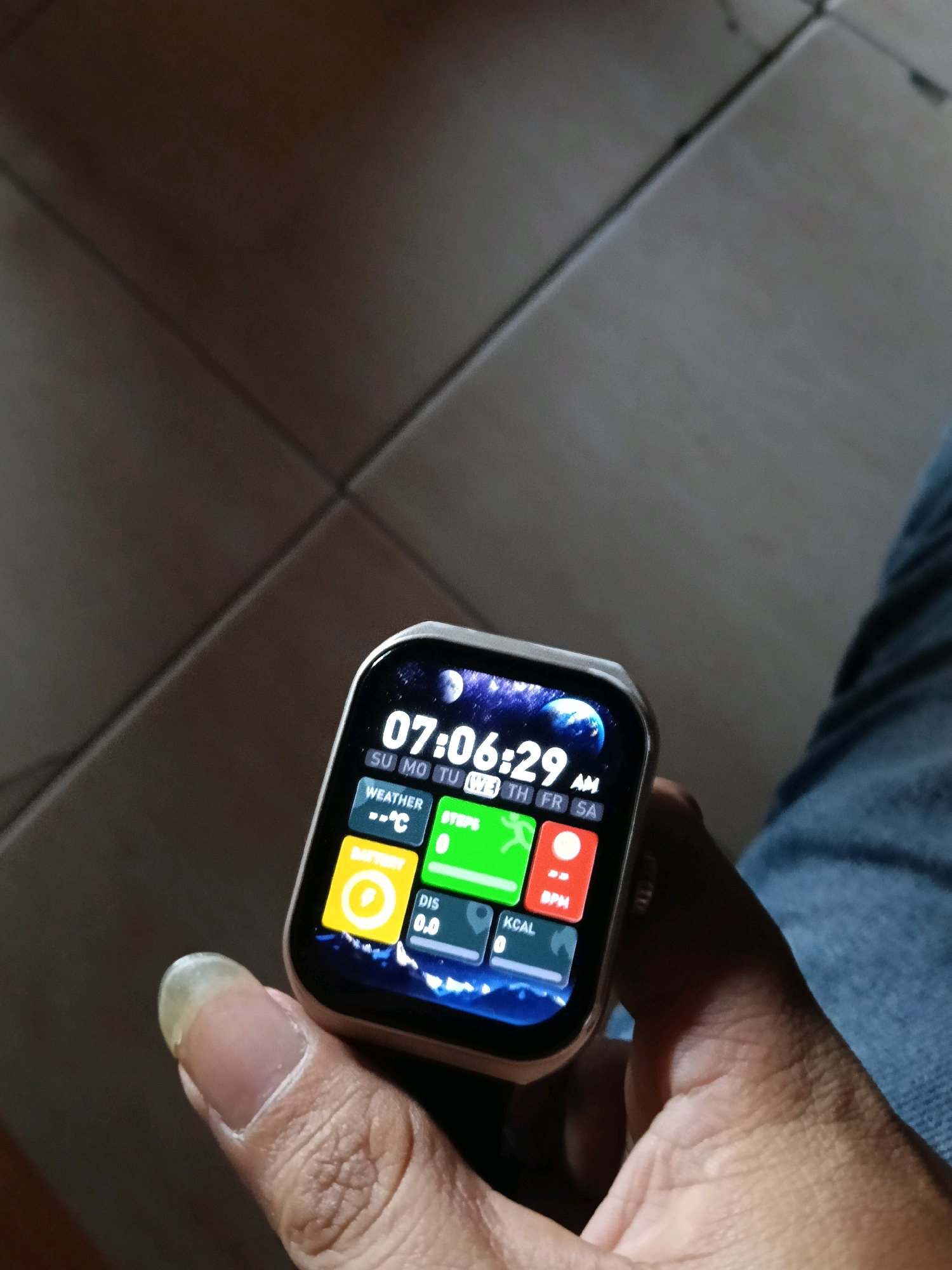 In Galuk, Indonesia, a person's hand, with a prominent fingernail, holds a smartwatch. The smartwatch displays the time as 07:06:29 AM, along with information about the weather, steps taken (0), distance covered (0.0), and BPM (heart rate). The background consists of a tiled floor.