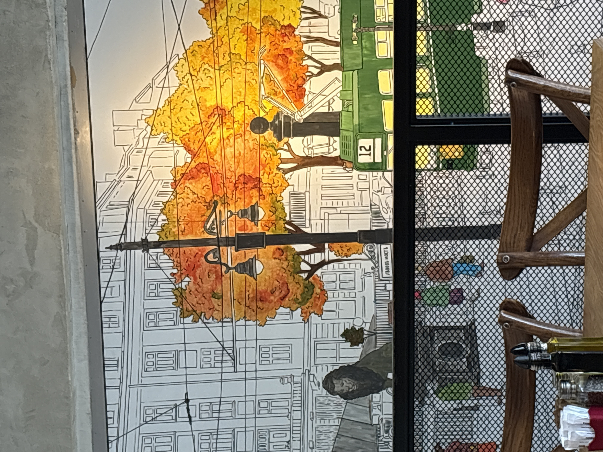 The image shows a mural depicting an urban scene. A green tram numbered "12" is prominently featured, surrounded by trees with autumn foliage in shades of yellow and orange. The background consists of a line drawing of city buildings, with lamp posts and utility lines adding to the urban ambiance. A female figure with long dark hair is seen near a sidewalk. The mural includes the words "Львов Мость". A chair and a metallic net rack partially obscure the right side of the mural, with items such as bottles and papers visible within the rack. The overall location appears to be an interior space, with a concrete column seen on the left edge of the image.