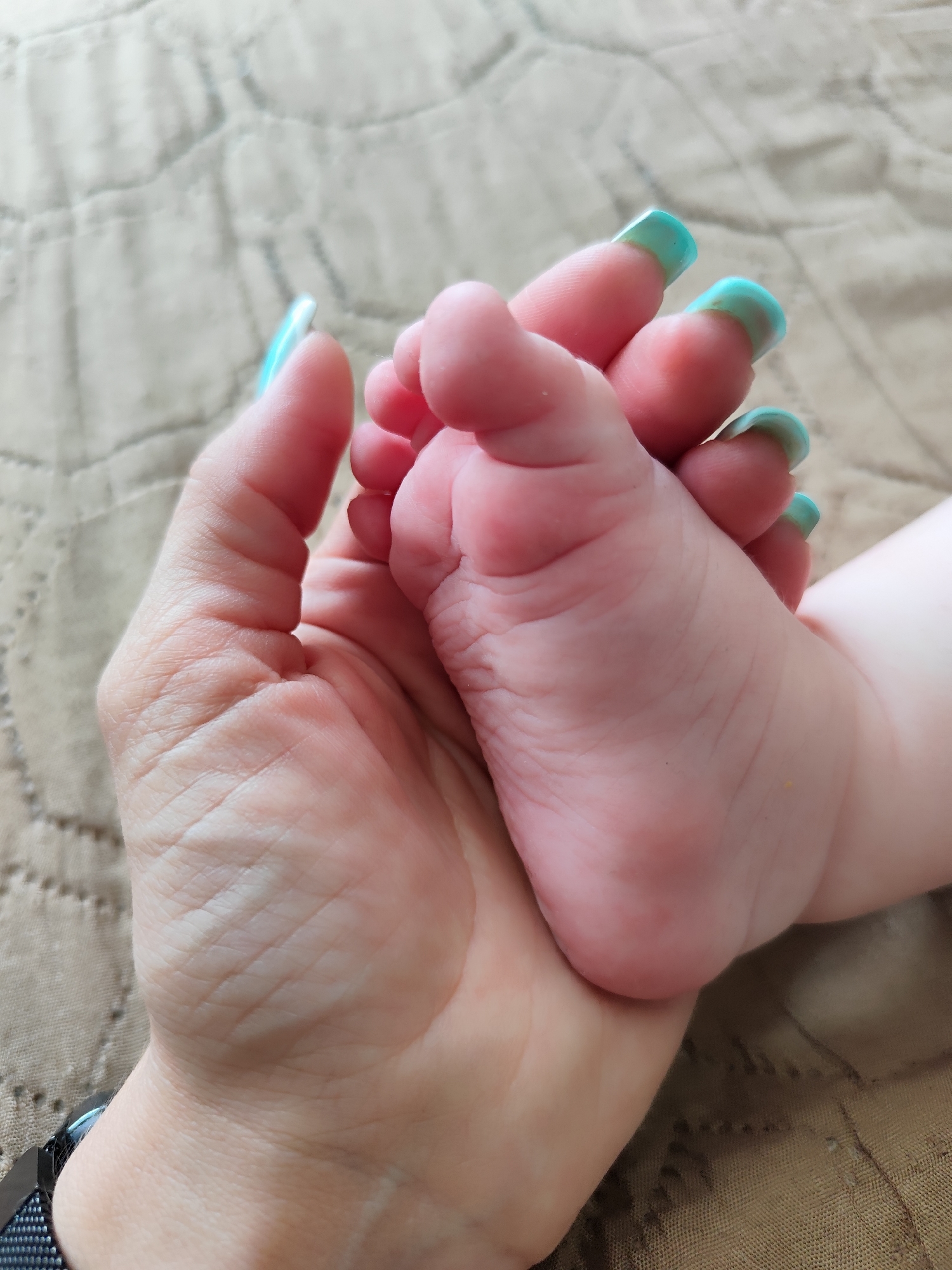 A baby's foot is held in a woman's hand. The woman has long, turquoise fingernails. The background is a textured beige surface. No location in the city of Фоки, Russia is visible or deducible from the image.