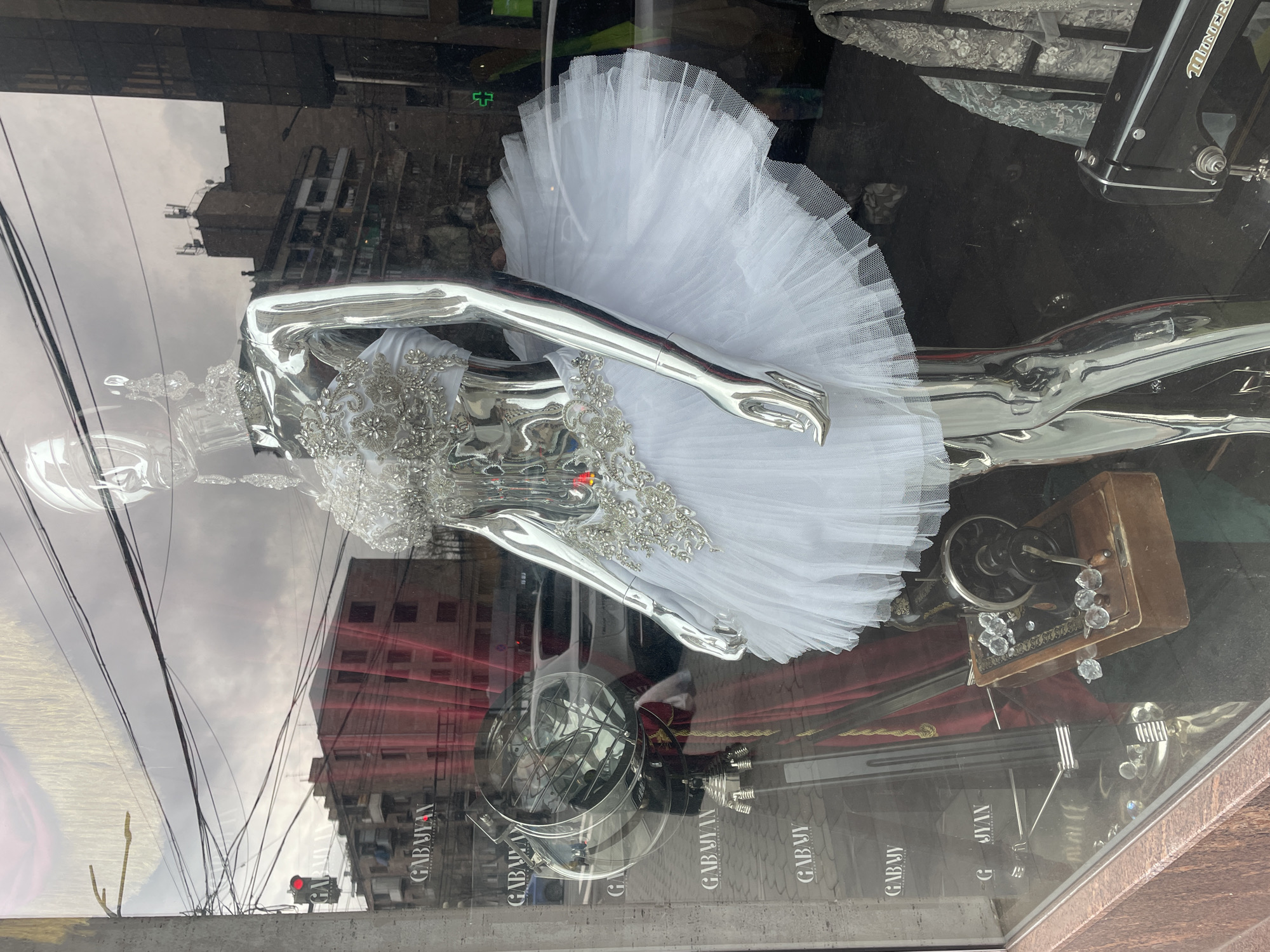 A shiny, chrome-colored mannequin torso with a white tulle skirt and silver-colored detailing is on display behind a glass window in a shop. The name “GABYUAN” is displayed several times on the glass of the window. A vintage sewing machine is on display behind the mannequin. Reflections visible on the glass show a street scene including a building with a red wall and a traffic light, which indicates the store may be in the city of Yerevan, Armenia.