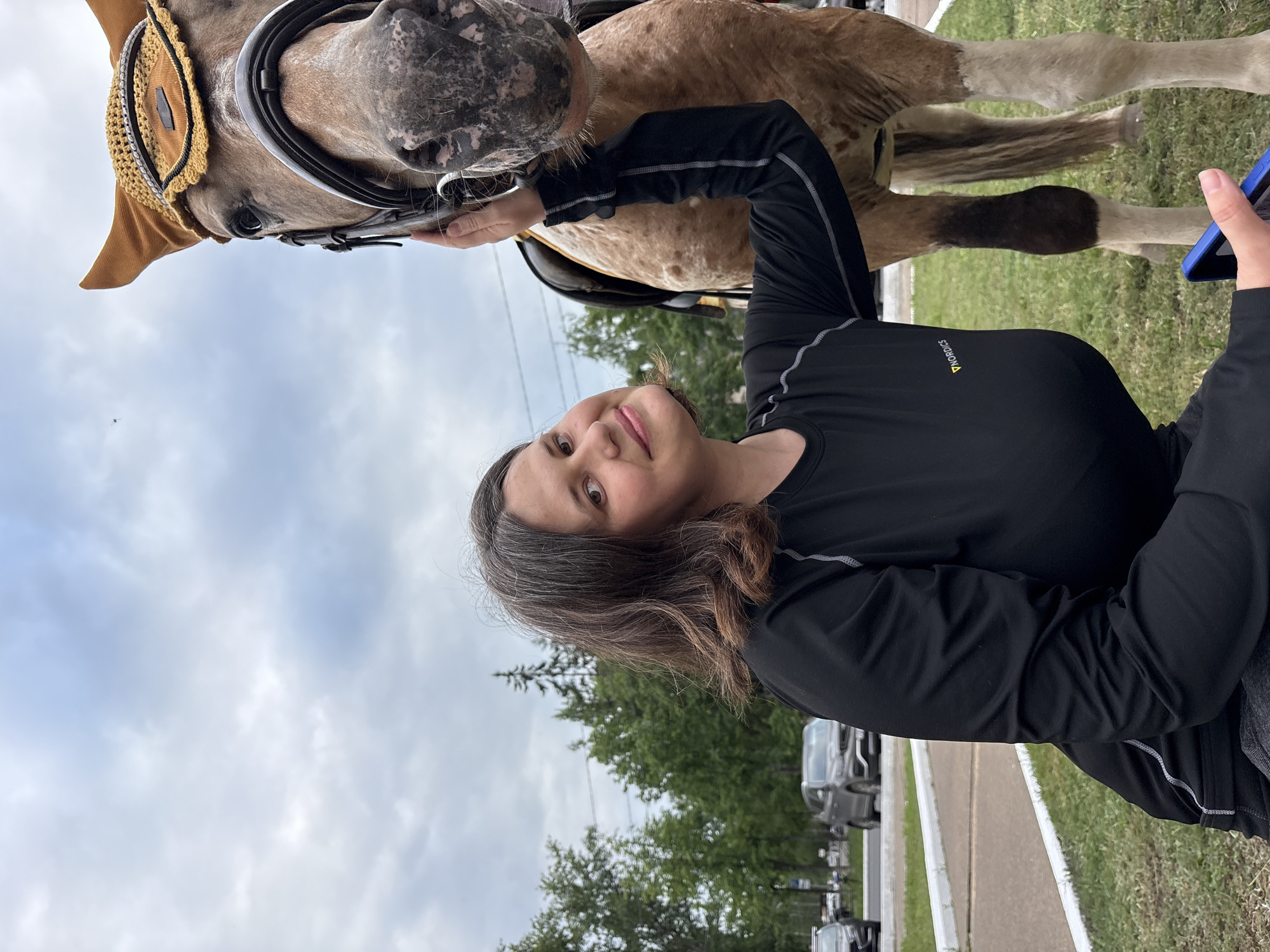 A woman is taking a selfie with a horse. The woman is wearing a black long-sleeved shirt and grey pants. She has long, dark brown hair with some grey streaks. The horse is light brown with a white muzzle and a brown bridle. The horse's head is close to the woman's face, and it appears to be nuzzling her. The woman is smiling at the camera.
In the background, there are trees and a cloudy sky. There are also some cars visible, suggesting a parking lot or a road nearby. The woman is holding a blue smartphone in her right hand, which is positioned to take the photo. The overall impression is a casual and friendly interaction between the woman and the horse. The lighting suggests it is daytime, with overcast weather.