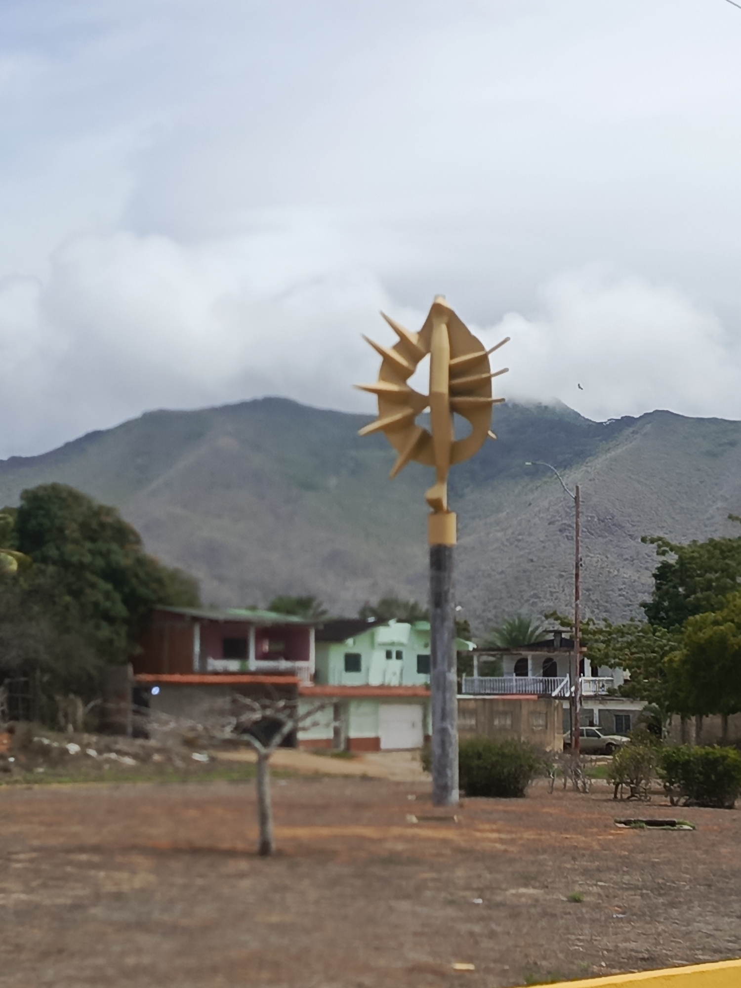 This is a landscape image captured in Pampatar, Venezuela, depicting an outdoor scene under an overcast sky. In the mid-ground, slightly right of center, a prominent golden, abstract sculpture stands atop a weathered grey pole. The sculpture features a central ovoid element from which multiple sharp, blade-like forms radiate outwards.
The immediate foreground consists of dry, reddish-brown earth or gravel, with sparse, low vegetation and a single thin, dark, bare tree trunk positioned left of the sculpture. A yellow line or curb is visible along the bottom edge of the frame.
Behind the sculpture and to its left, several buildings are visible. One notable structure is a two-story building with a reddish-brown upper floor, white railings, and a balcony, situated above a lighter-colored ground floor. Adjacent to it on the right is a two-story building with a light green facade. To the right of the sculpture, a wooden utility pole with a street light and overhead wires is present. A light-colored sedan is partially visible behind some bushes and another building further back on the right.
The background is dominated by a range of large, undulating mountains, appearing greyish-green due to sparse vegetation or rock. Clouds partially obscure the mountain peaks, and a small, dark bird is discernible flying against the sky above the mountains, to the right of the sculpture. The sky is predominantly grey with patches of white clouds, indicating an overcast day. No people are visible in the scene.