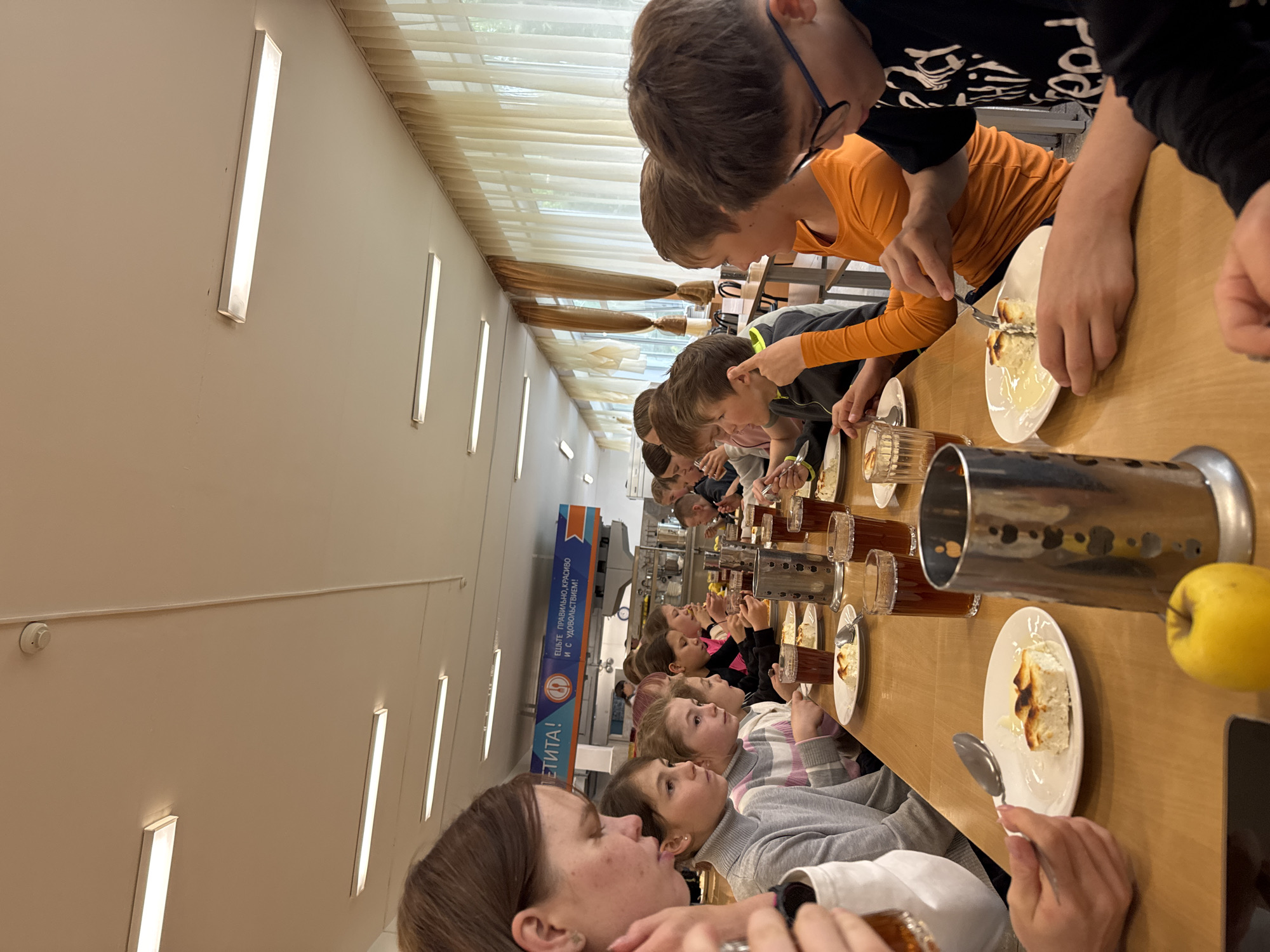 This rotated image captures an indoor dining scene, likely a school cafeteria or dining hall, in Chaikovsky, Russia. A long line of children, appearing to be elementary to early teen students, are seated at a wooden table, focused on their meal.
Each child has a white plate containing a portion of a baked dessert, possibly a cheesecake or sweet casserole with a lightly browned crust, often served with a light-colored sauce or syrup. Glasses filled with a dark beverage, such as tea or juice, are also present on the table. In the foreground, a bright yellow apple sits on the table next to a metal utensil holder. The children are actively eating, holding forks and spoons.
The room is brightly illuminated by both natural light filtering through large windows covered with cream-colored blinds and by numerous rectangular LED light fixtures embedded in the ceiling and walls. In the background, a blue and orange banner is affixed to the wall. The text on the banner is in Russian and reads: "Приятного АППЕТИТА!" (Enjoy your meal!) and "ЕШЬТЕ ПРАВИЛЬНО, КРАСИВО И С УДОВОЛЬСТВИЕМ!" (Eat correctly, beautifully, and with pleasure!). The scene depicts an orderly mealtime, with children enjoying their food in a well-lit environment.