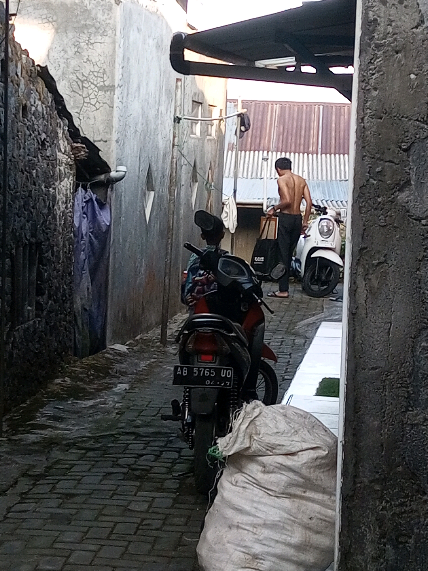 The image depicts a narrow alleyway with weathered concrete walls and a cobblestone ground. On the left, a stone wall is partially covered by a large purple tarp. A motorcycle is parked in the foreground, with its license plate reading "AB 5765 UO". In the background, a shirtless man is walking away from the camera, carrying a tote bag and a brown box. Another motorcycle, a white scooter, is parked behind him. The scene appears to be in Andongsili, Indonesia, suggesting a typical urban environment with residential structures and common modes of transportation. The lighting indicates it is daytime, possibly late afternoon or early morning, given the shadows. The overall impression is one of a functional, lived-in space, possibly a backstreet or service alley.