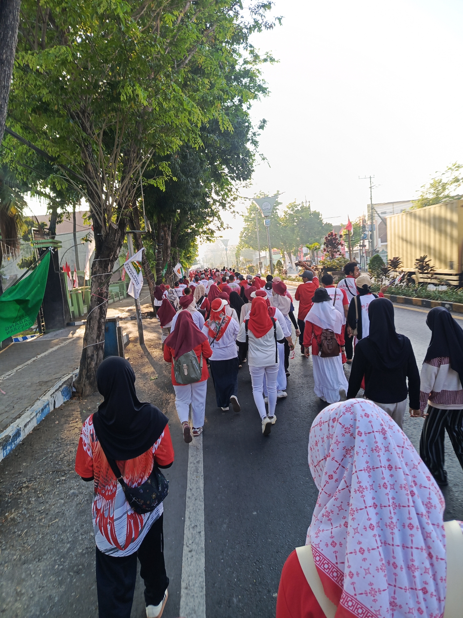 In Semampir, Indonesia, a group of people are walking down a paved street lined with trees. The group appears to be participating in an organized event, with many individuals wearing matching red and white attire, including headscarves and shirts. Some are carrying small bags or wearing backpacks. The street is also decorated with flags, some of which are red and white. In the background, there are buildings, a large yellow shipping container, and power lines. The sky is bright, suggesting it is daytime. The scene depicts a communal activity taking place in an urban environment.