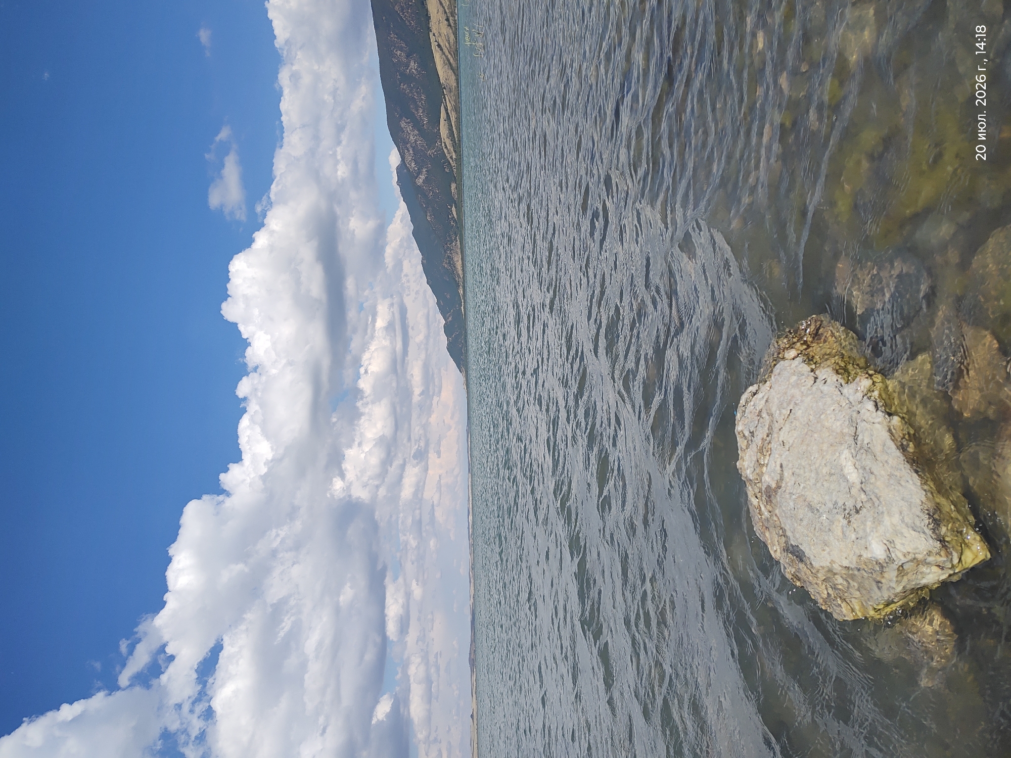 The image depicts a wide body of water, likely a lake, under a bright blue sky with scattered white clouds. In the foreground, a large, light-colored rock sits partially submerged in the clear water. The water exhibits ripples, suggesting a gentle breeze. In the distance, a chain of dark, sloping hills or mountains forms the horizon, with some sparse vegetation visible on their slopes. The overall scene is natural and serene, with no human presence or man-made structures immediately apparent. A timestamp in the bottom right corner reads "20 июл. 2026 г., 14:18", indicating the date and time the photograph was taken. The location of Акылбай, Kazakhstan is not visually discernible from the image itself, as it is a general landscape scene.