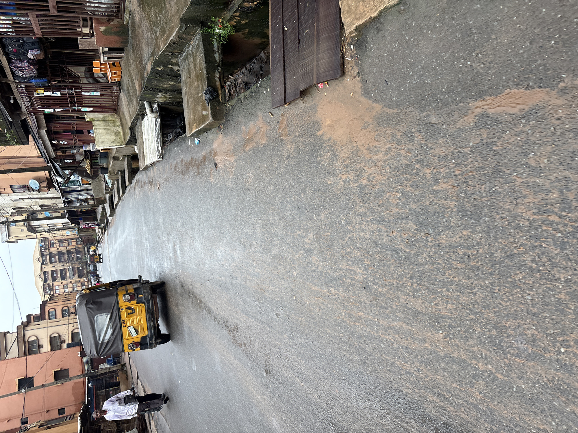 The image depicts a street scene in Onitsha, Nigeria. A yellow auto-rickshaw, identified as a "TVS" model, is traveling away from the camera on a wet, asphalt road. To the left of the auto-rickshaw, a man in a white shirt and dark pants walks towards the camera, carrying a bag. The street is lined with buildings and shops, many with metal grates and awnings. A drainage system runs alongside the road, with visible concrete channels and a wooden plank serving as a makeshift bridge over one section. Muddy water is flowing in the drainage and has spread onto the road. The overall environment suggests an urban setting, possibly after rainfall, with signs of everyday life and infrastructure.
