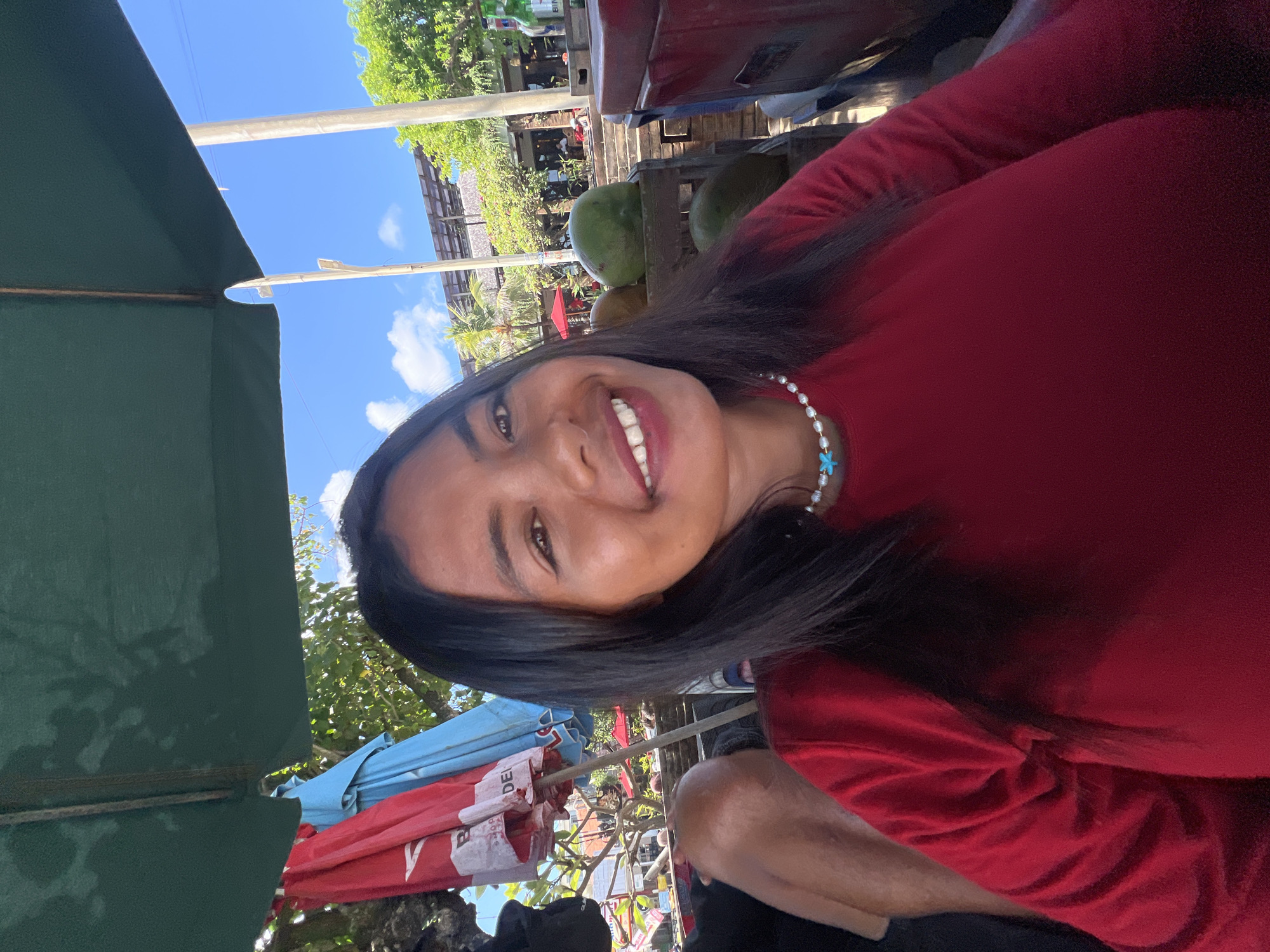 A young woman with long, dark hair smiles at the camera, seated outdoors under a green umbrella. She wears a red long-sleeved shirt and a pearl necklace with a blue pendant. In the background, red and blue umbrellas are visible, along with lush green trees and buildings. Two green coconuts are on display in a wooden crate to her right, and a red cooler is partially visible. The scene suggests a casual, sunny day in Kuta, Indonesia, likely at a beachfront cafe or market.