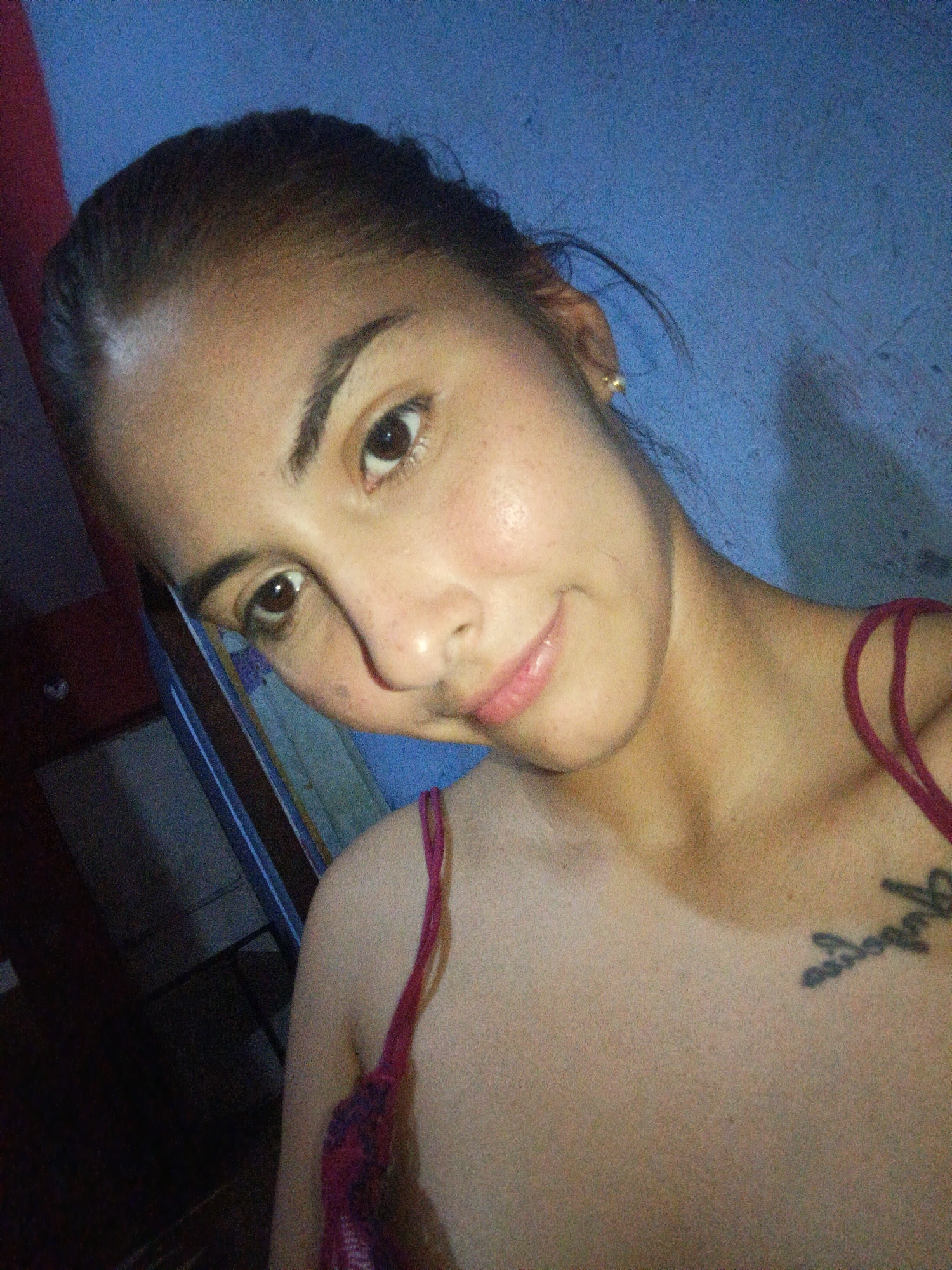 A medium-shot selfie taken indoors shows a light-skinned woman with dark hair pulled back, wearing a red tank top with spaghetti straps. She is positioned at an angle, facing the camera with a slight smile. A visible tattoo in dark ink is located on her upper right chest. The background consists of a blue wall. A dark cabinet or piece of furniture is partially visible in the lower left corner, with light-colored framework visible. The location of Juan Griego, Venezuela is not evident in the image.