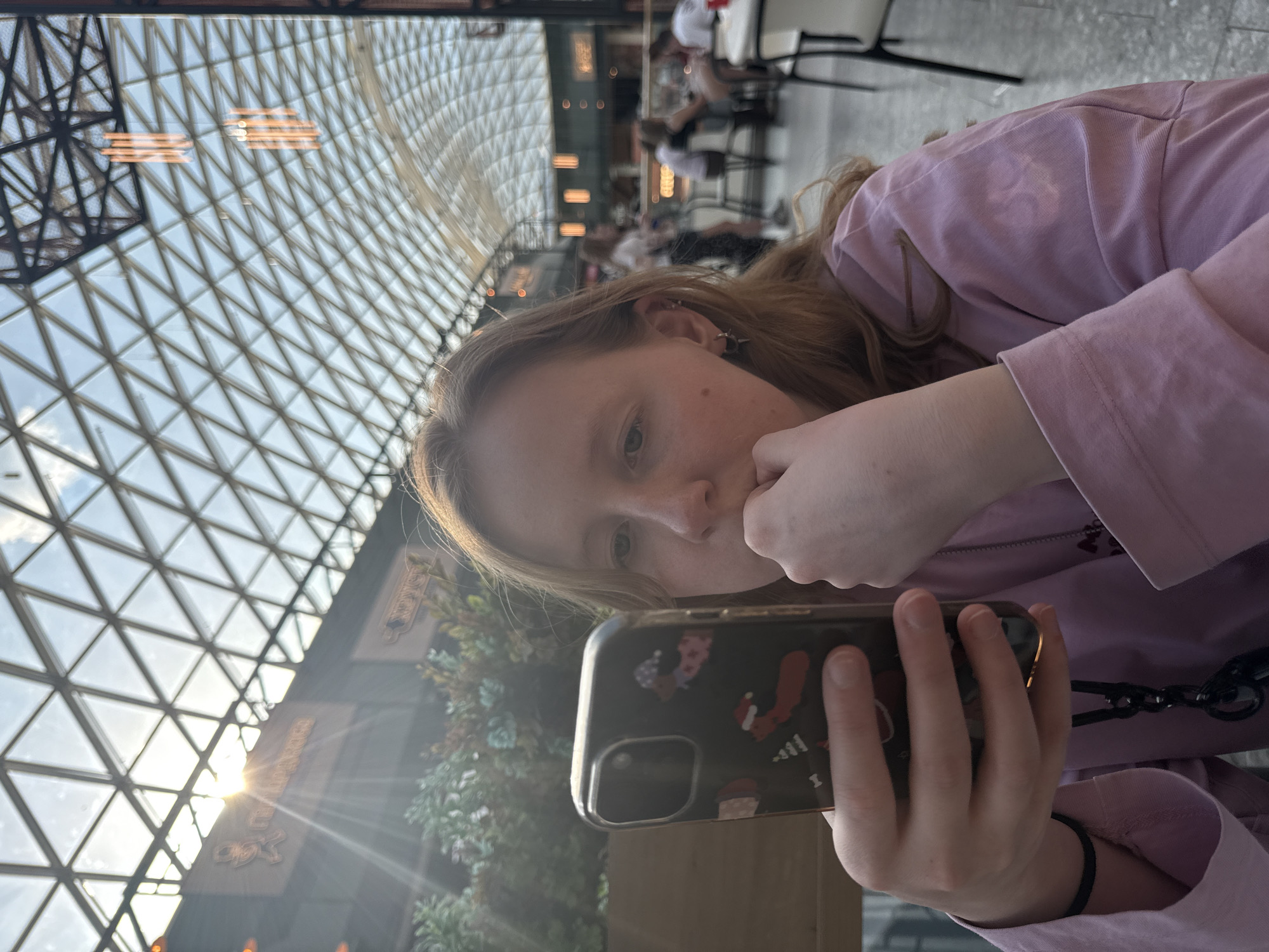A young woman is shown from the chest up, angled downward and to the left, holding a smartphone with a dark case with a cartoon design. She is wearing a light purple top and resting her chin on her hand. The background reveals an interior space with a high, gridded ceiling made of glass and metal. A blurry scene of people and tables suggests a cafe or public area. Light streams in from the left side of the image. Based on the style of architecture and interior, the location could be the Planeta Shopping Mall in Perm, Russia.