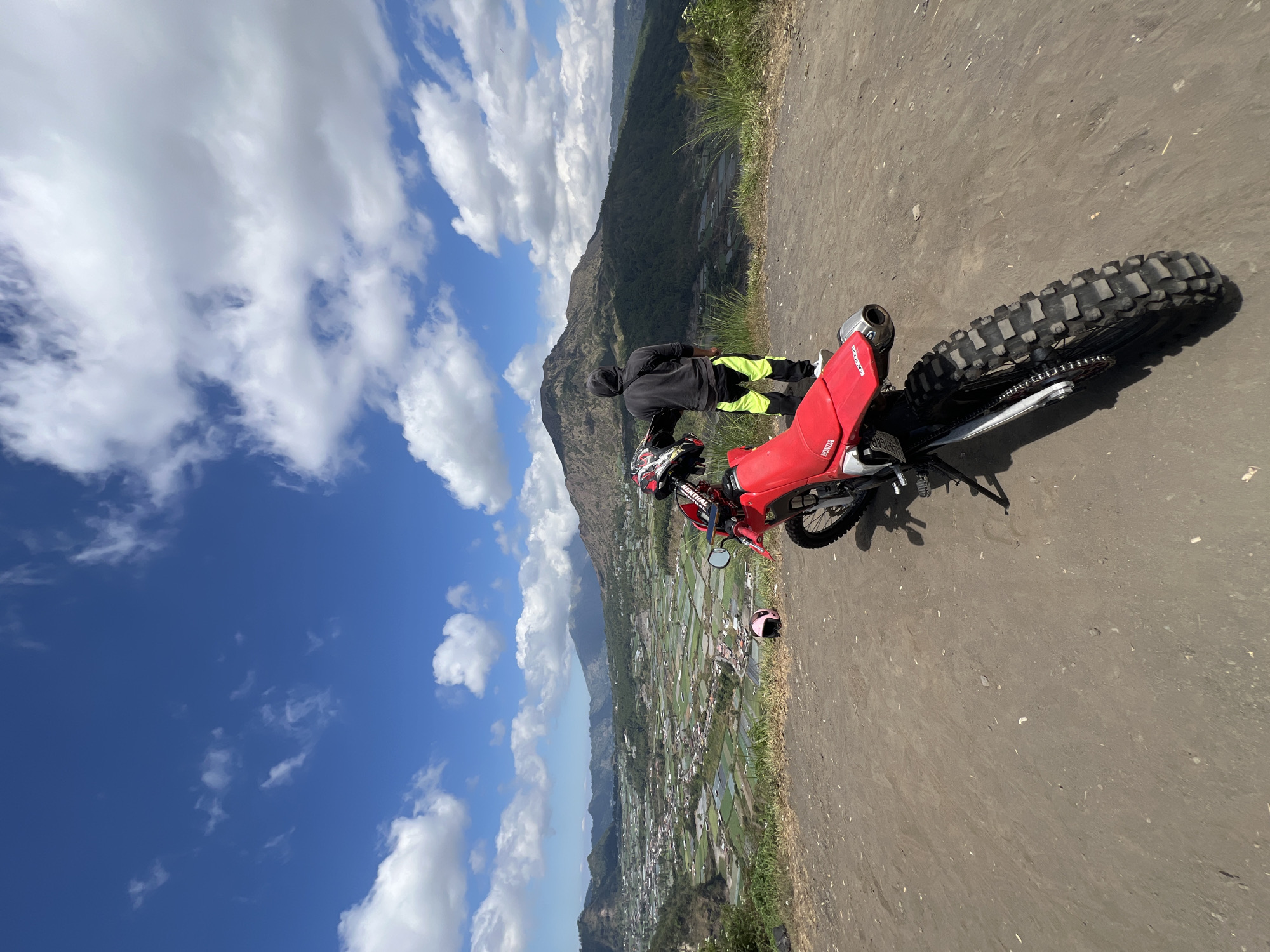 A person stands on a dirt path overlooking a valley in Kintamani, Indonesia. A red dirt bike is parked in the foreground, with two helmets resting nearby. The person, wearing a dark hoodie and bright yellow pants, faces away from the camera, gazing at the vast landscape. In the distance, rolling hills and a town are visible, with a prominent mountain in the background. The sky is a vibrant blue, dotted with fluffy white clouds, indicating a clear day. The terrain is rugged and earthy, typical of a viewpoint in a mountainous region.